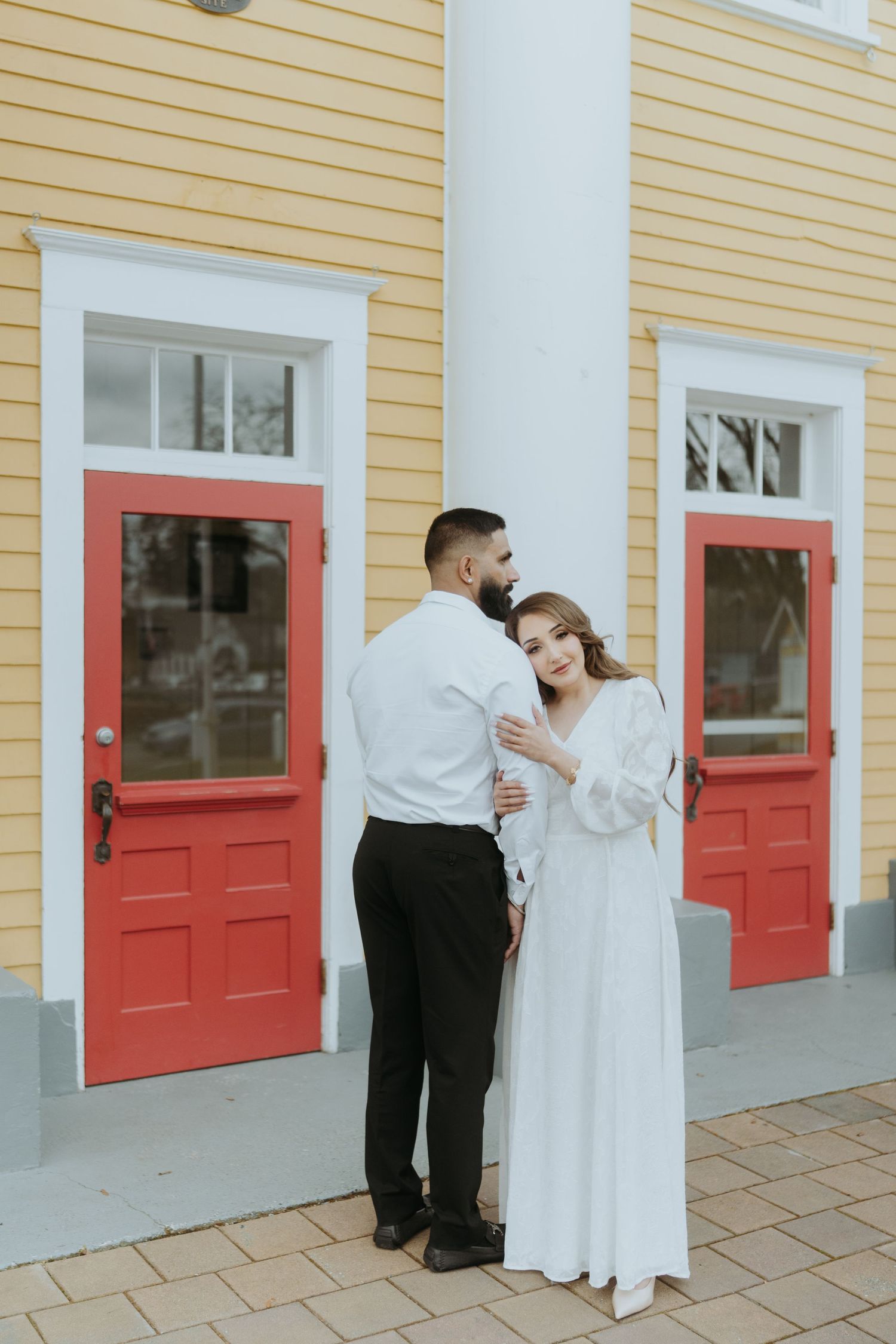 A couple poses for photos in front of a yellow building with bright red doors.
