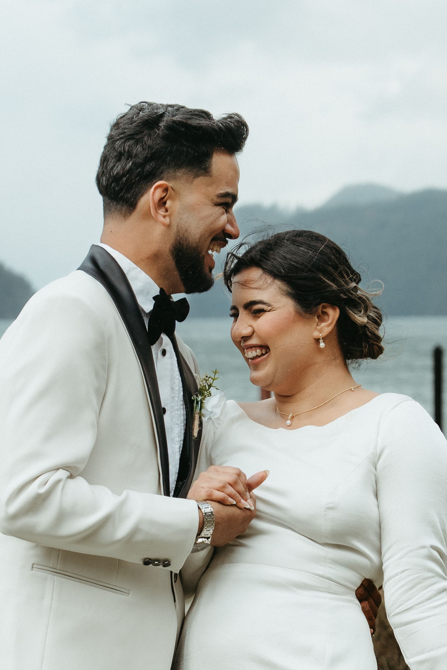 A couple in white wedding attire share joyful laughter together against a scenic mountain backdrop.