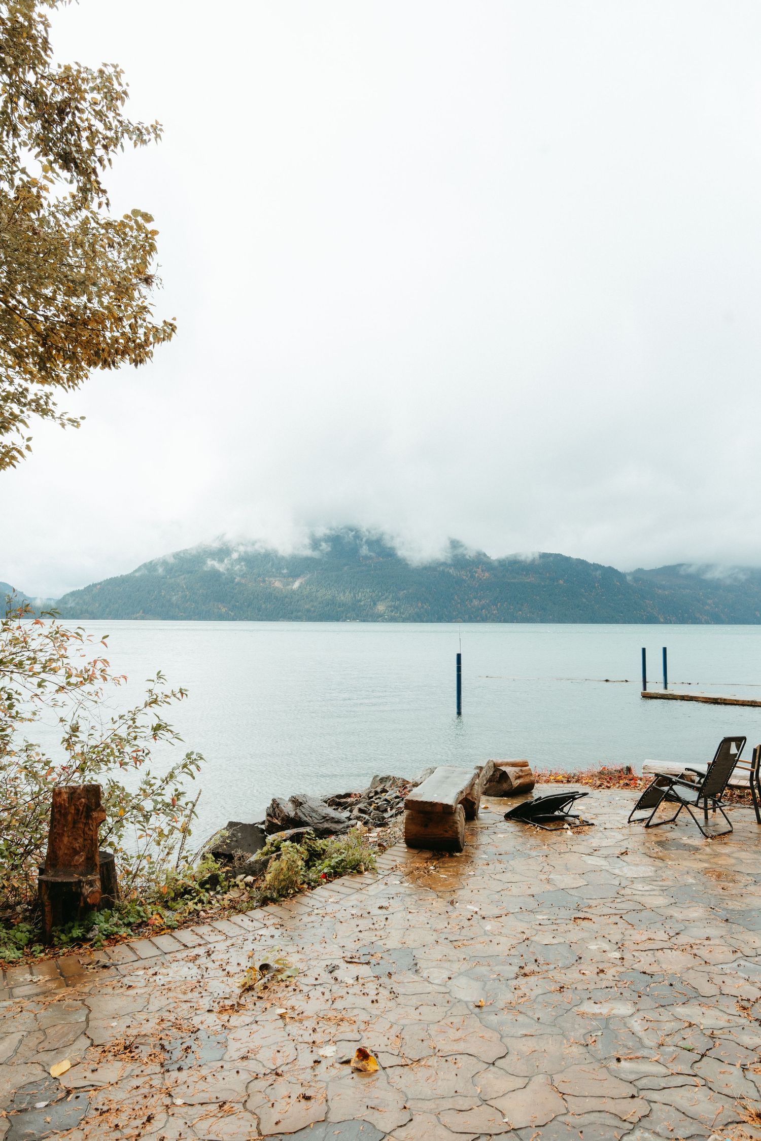 A misty lakeside stone patio with wooden chairs overlooking mountains and water on a foggy autumn day.