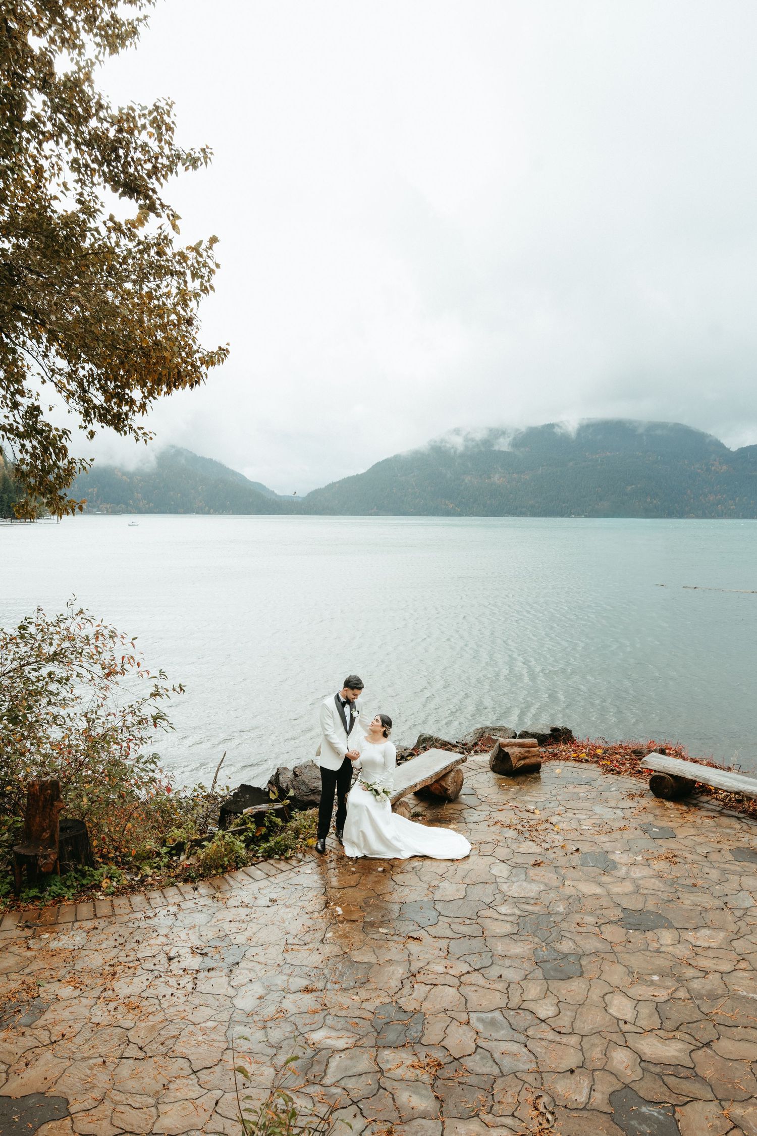A misty lakeside scene with stone patio overlooking mountains and water, featuring figures in white clothing.