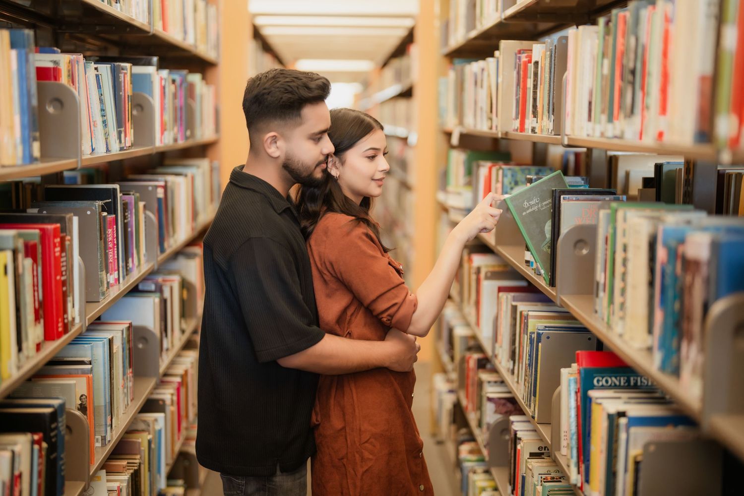 A couple embraces while browsing books in a library's cozy wooden shelves.