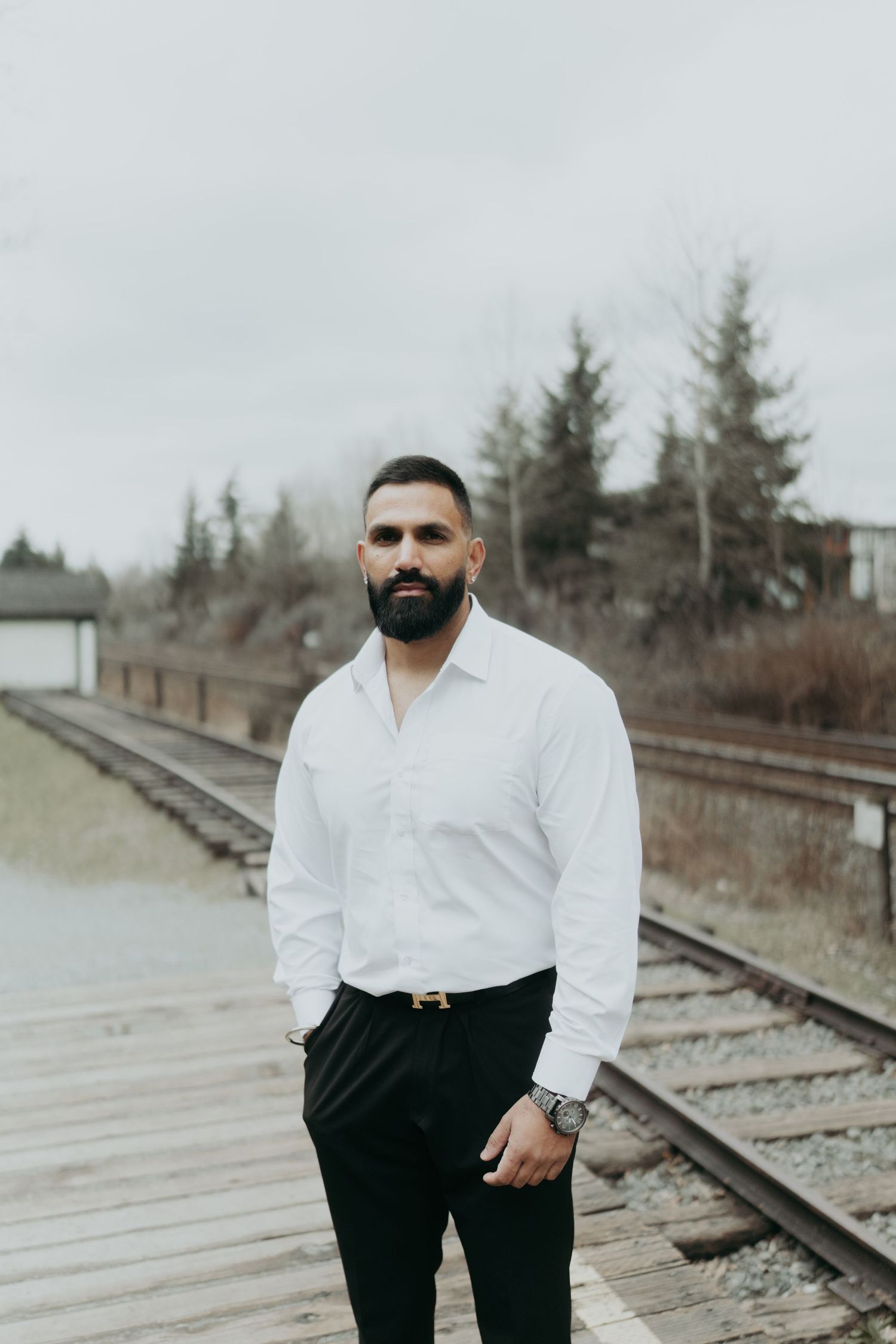 Person in white shirt and black pants poses near railroad tracks on an overcast day.