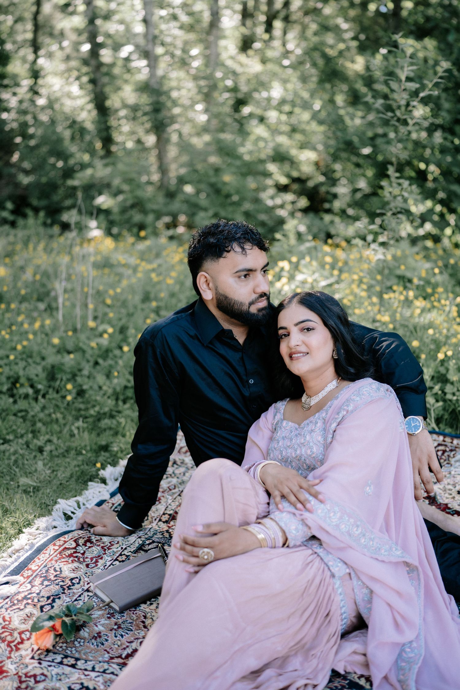 Couple lounges on patterned blanket in outdoor setting with pink dress and black attire.