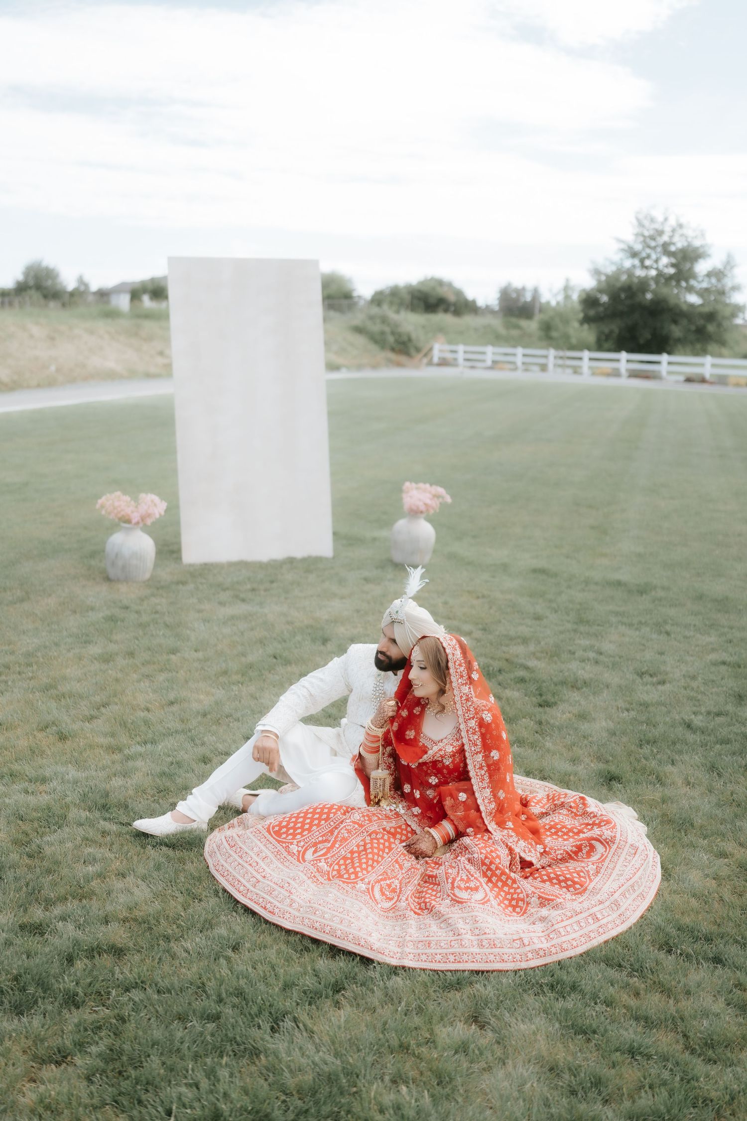 Couple in traditional Indian wedding attire sitting on grass, bride in red lehenga and groom in white sherwani.