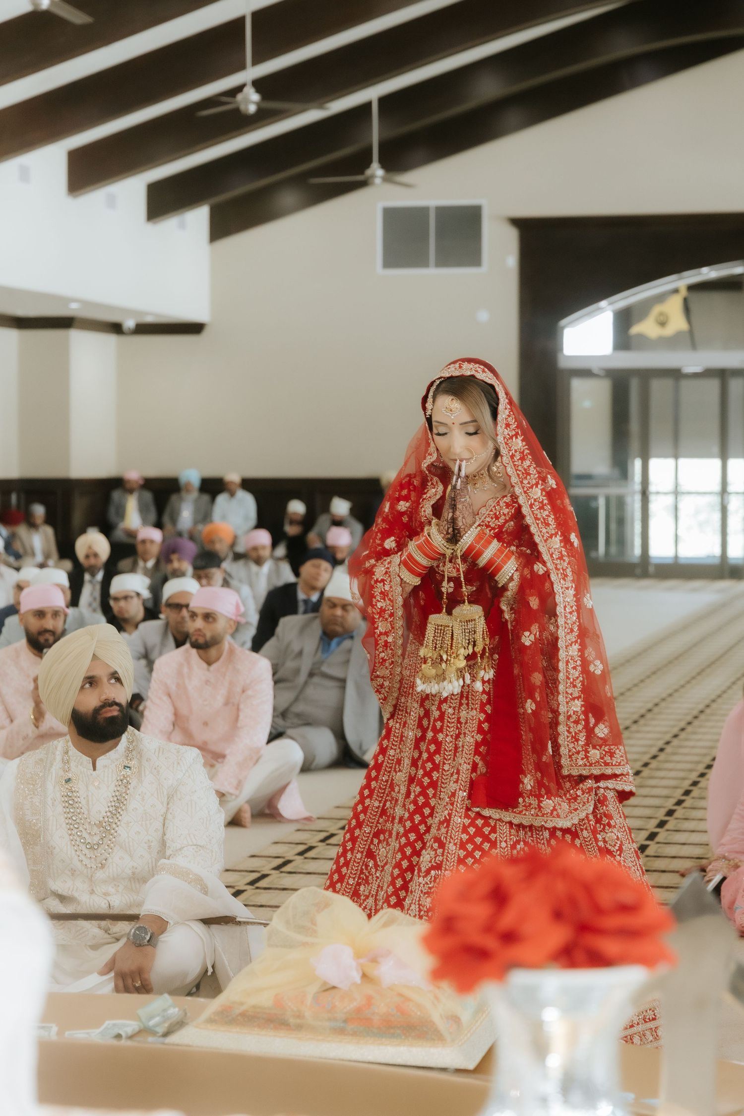 Sikh bride in red lehenga praying during Anand Karaj wedding ceremony at Gurudwara with guests seated.