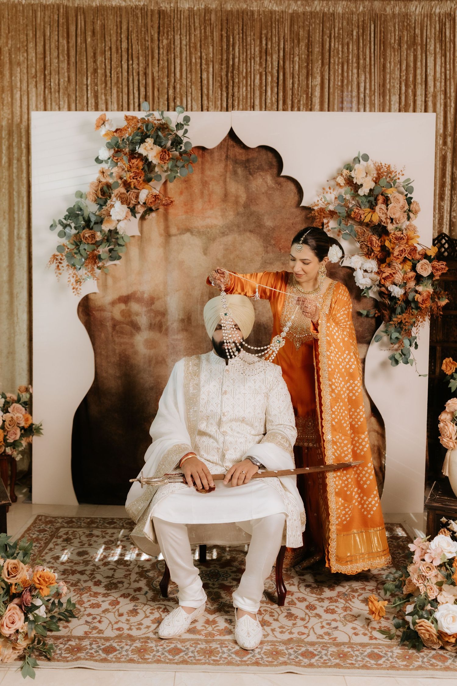 A bride in orange saree places a garland on groom in white sherwani at a floral South Asian wedding ceremony.
