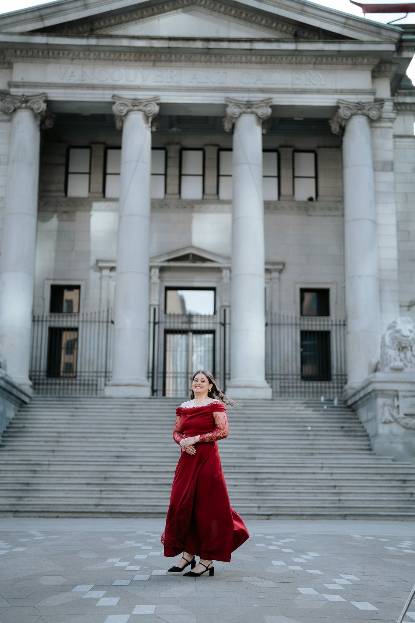 A woman in an elegant red off-shoulder gown poses in front of a grand neoclassical building with columns.