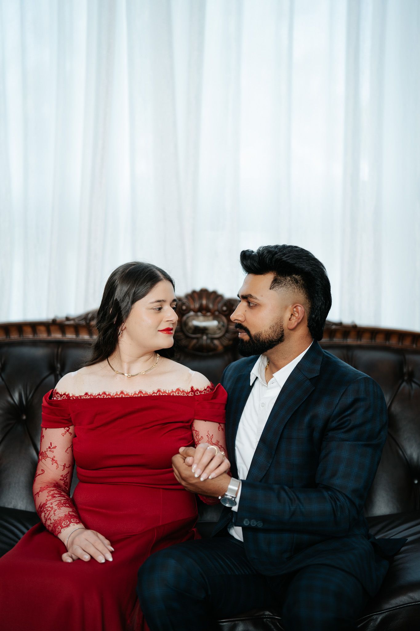Couple in elegant attire sitting on leather sofa, woman in red lace gown and man in dark suit holding hands.