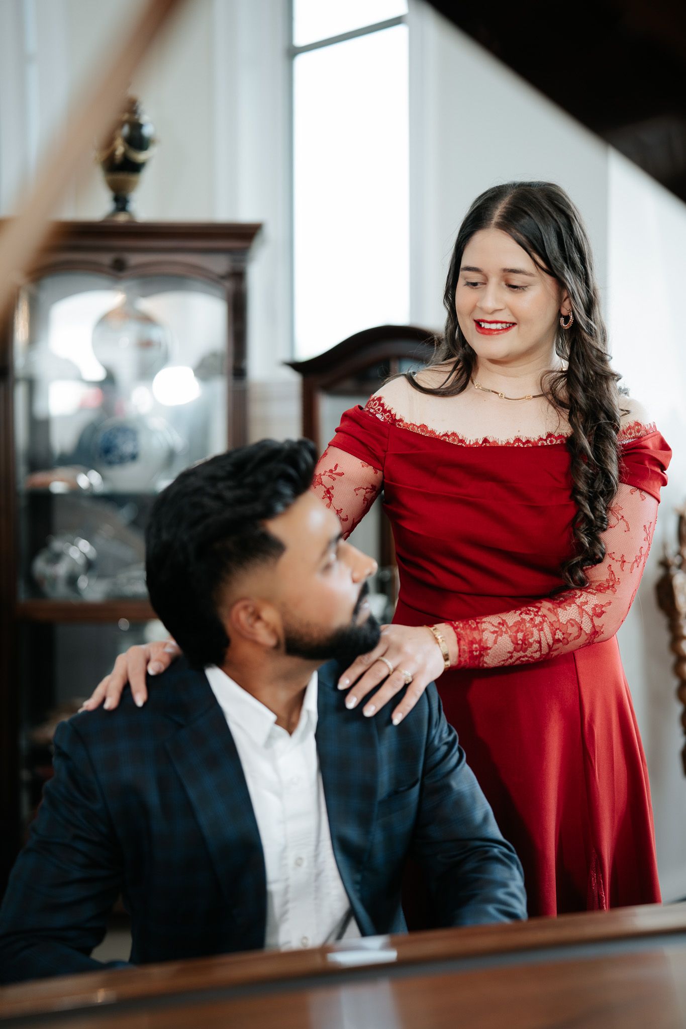 A woman in a red lace off-shoulder gown places her hand on a bearded man in a dark suit at a piano.