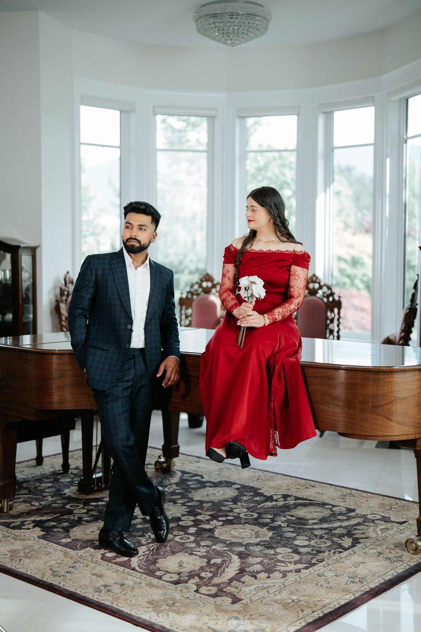 Couple posing in elegant white room with grand piano, woman in red lace gown holding white bouquet, man in plaid suit.