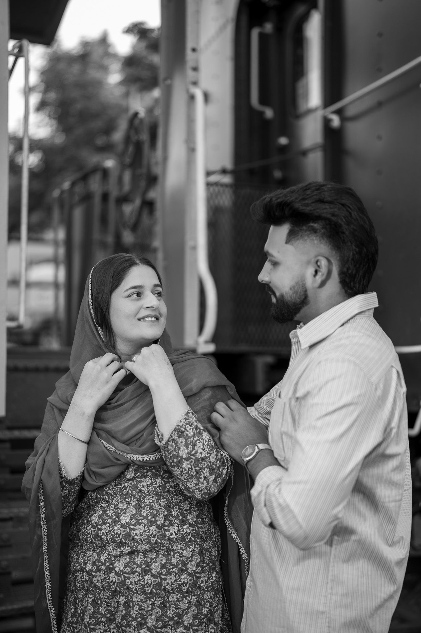 A couple shares a tender moment at a train station, the woman in a floral salwar and dupatta smiling at her bearded partner.