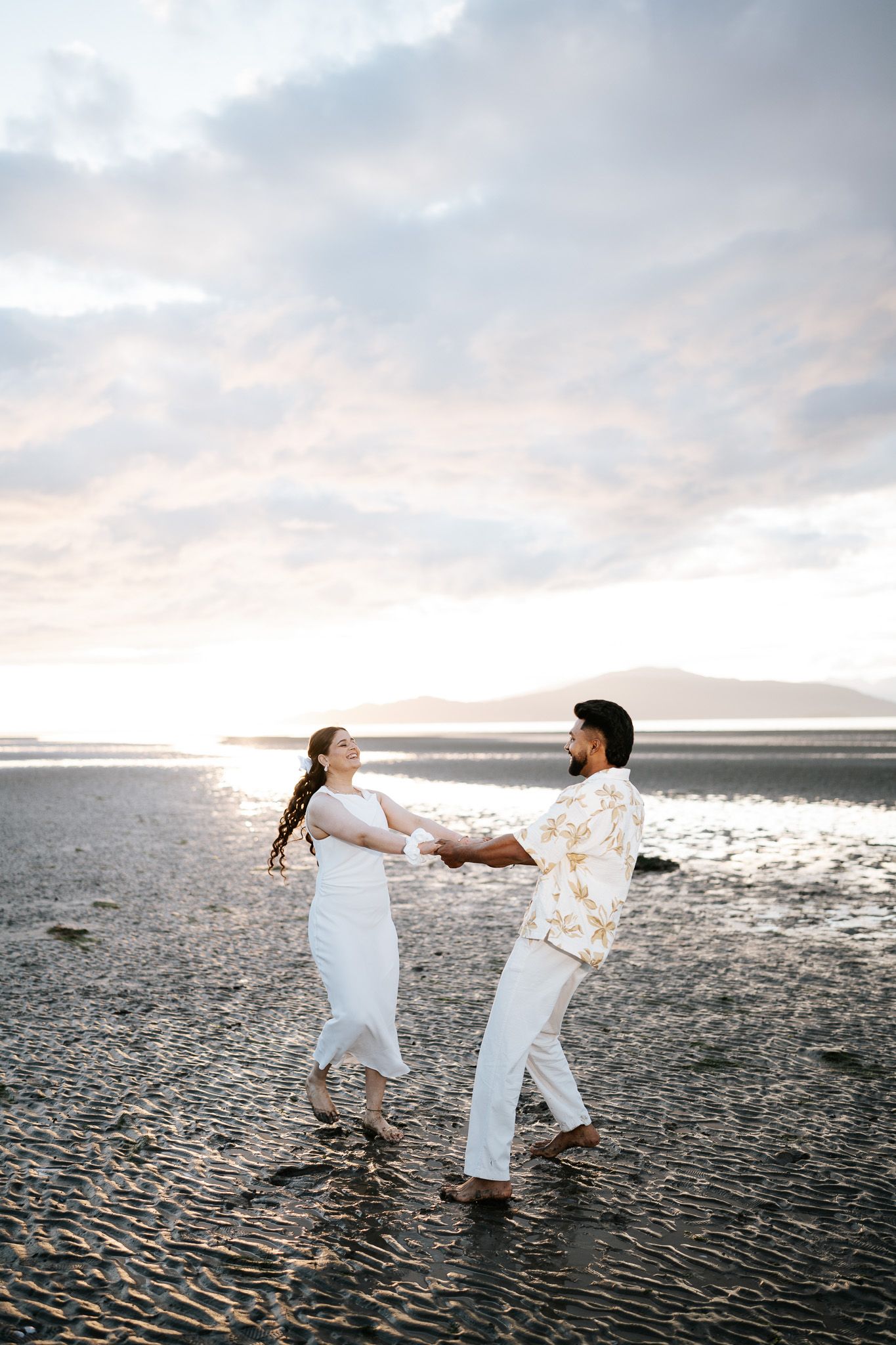 Couple holding hands dancing on a reflective beach at sunset, wearing white and floral outfits.