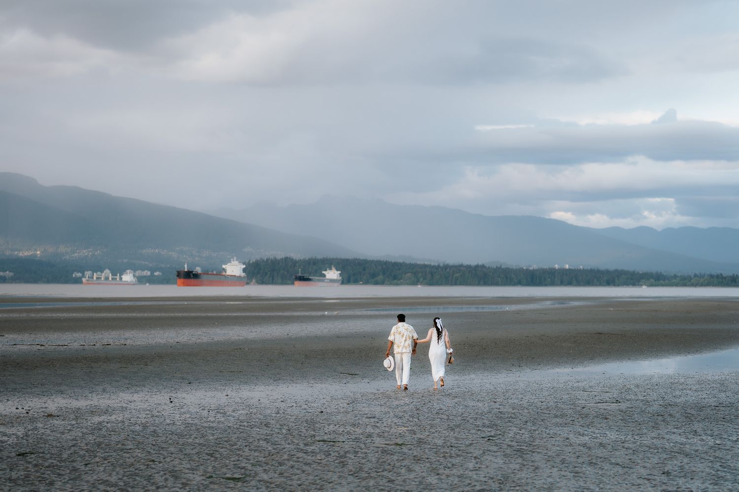 Couple in white walking on a vast tidal flat with a red cargo ship and misty mountains in the background.