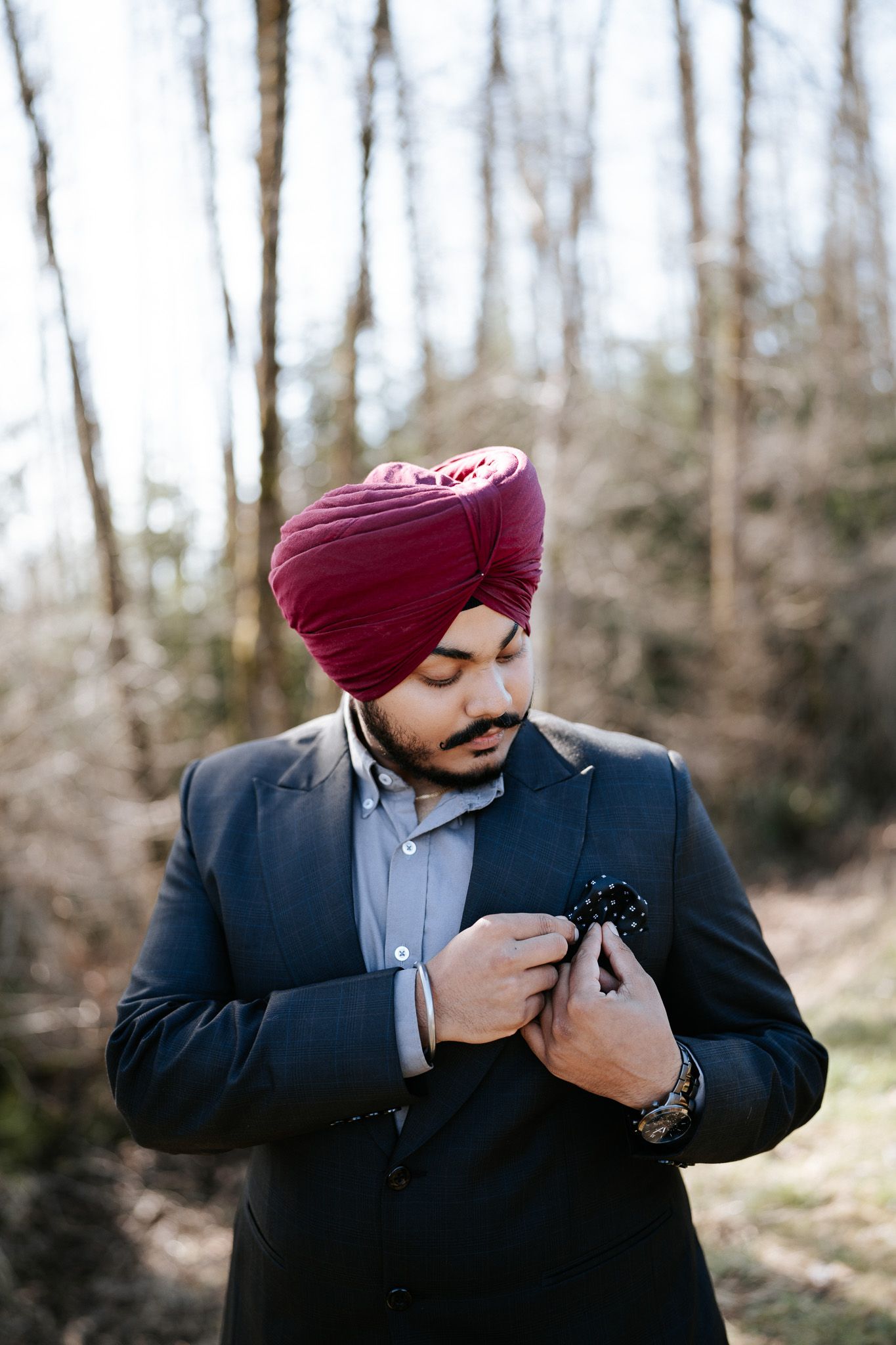 Person in a navy suit and burgundy turban adjusts their outfit while standing in a wooded outdoor setting.