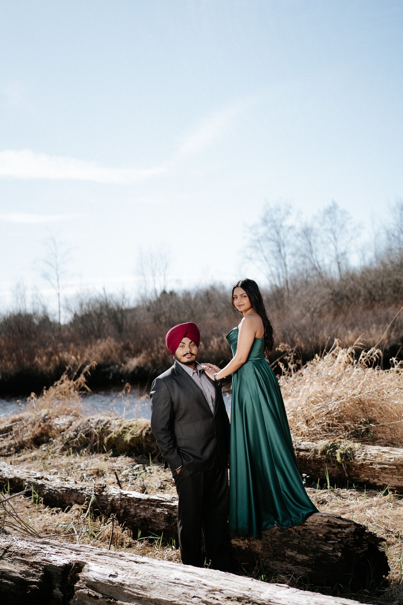 A couple in formal attire, with a green gown and dark suit, share an intimate moment in a winter field at sunset.