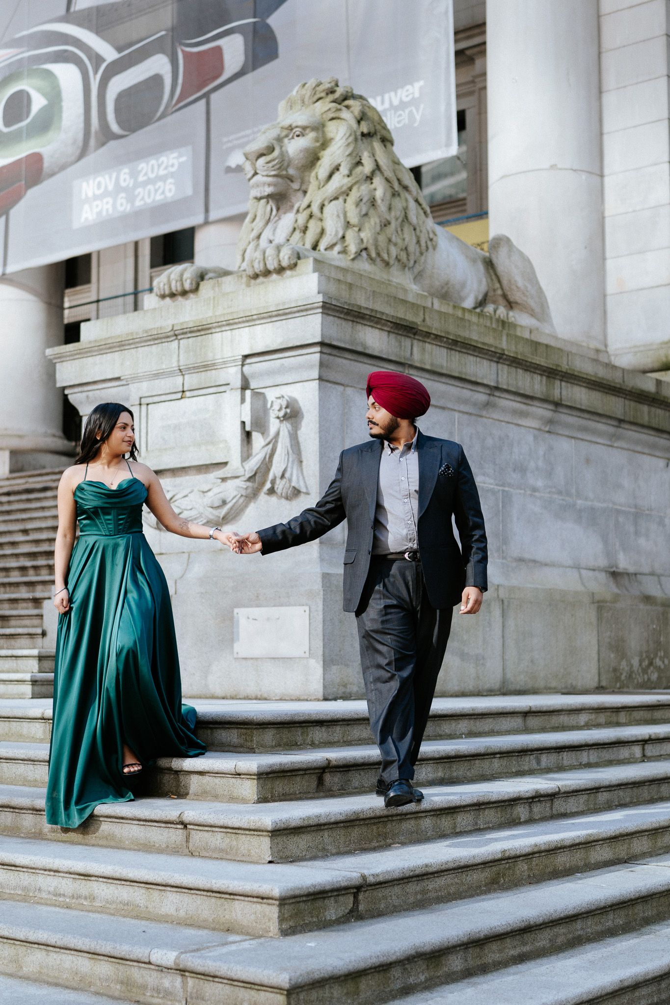 Couple in formal attire poses playfully on grand marble steps with stone lion statues at a historic building entrance.