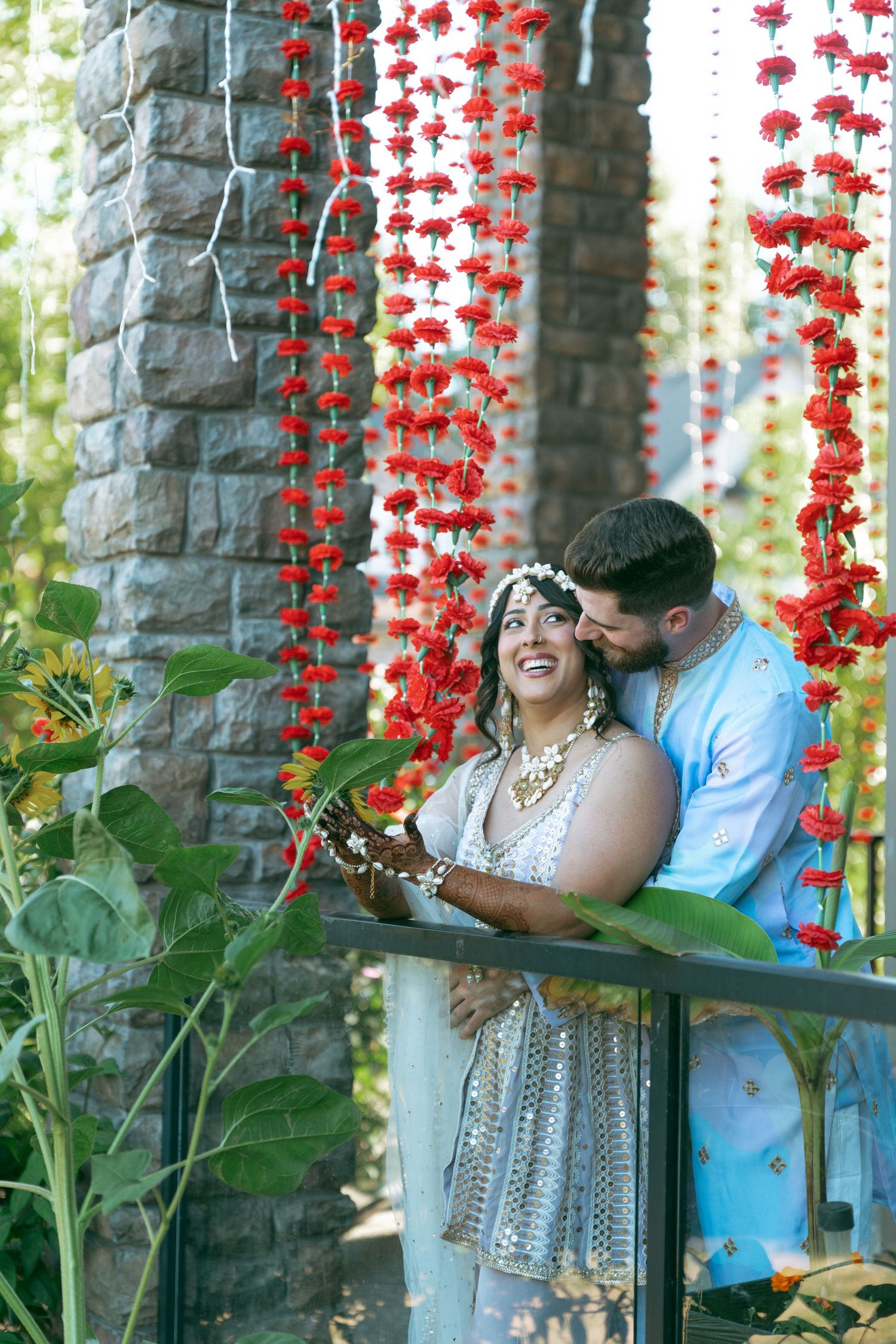Couple in traditional Indian wedding attire smiling under red floral garlands outdoors.