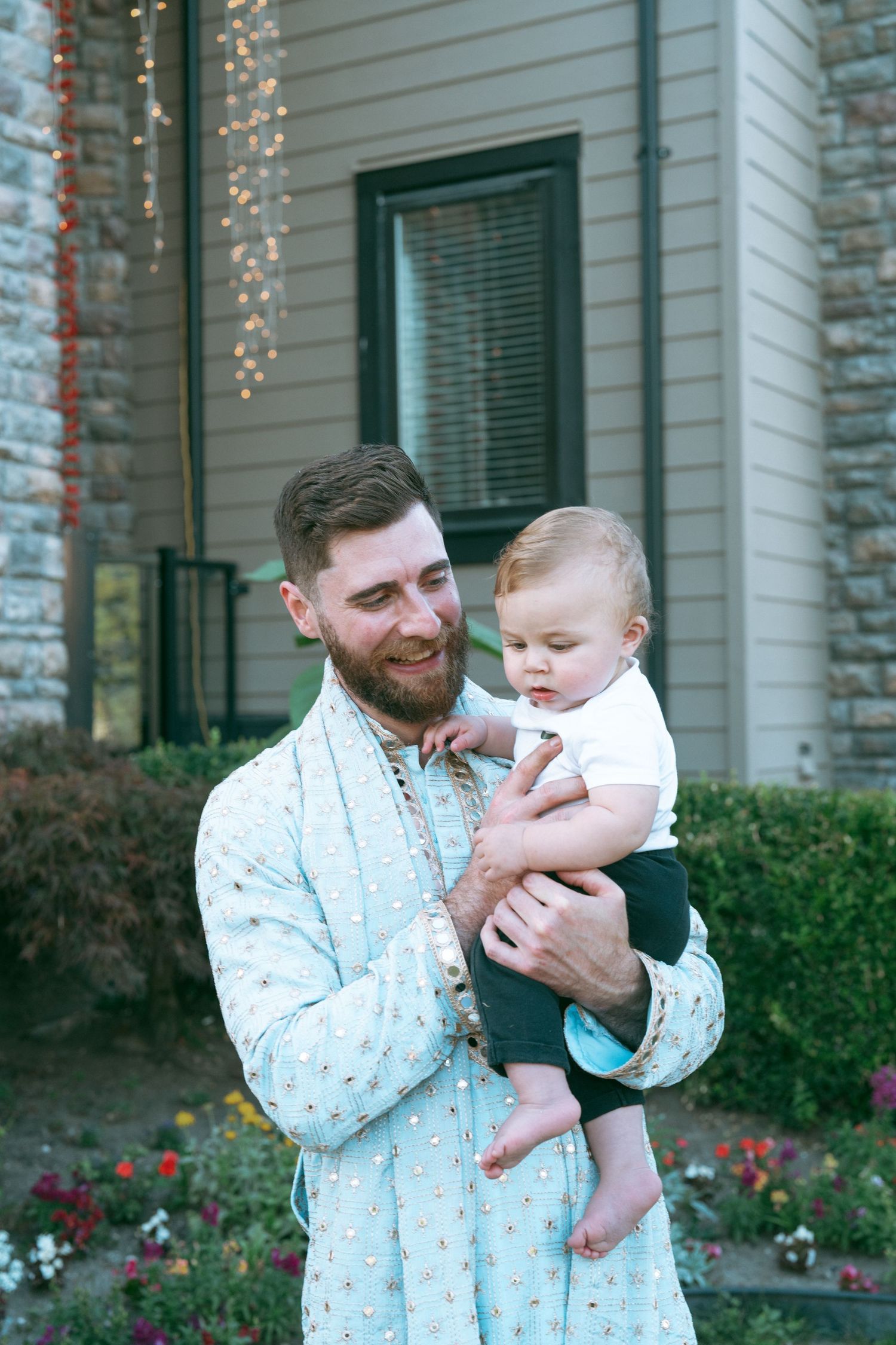 Bearded man in floral robe smiling while holding a baby boy outdoors in front of a house with string lights.