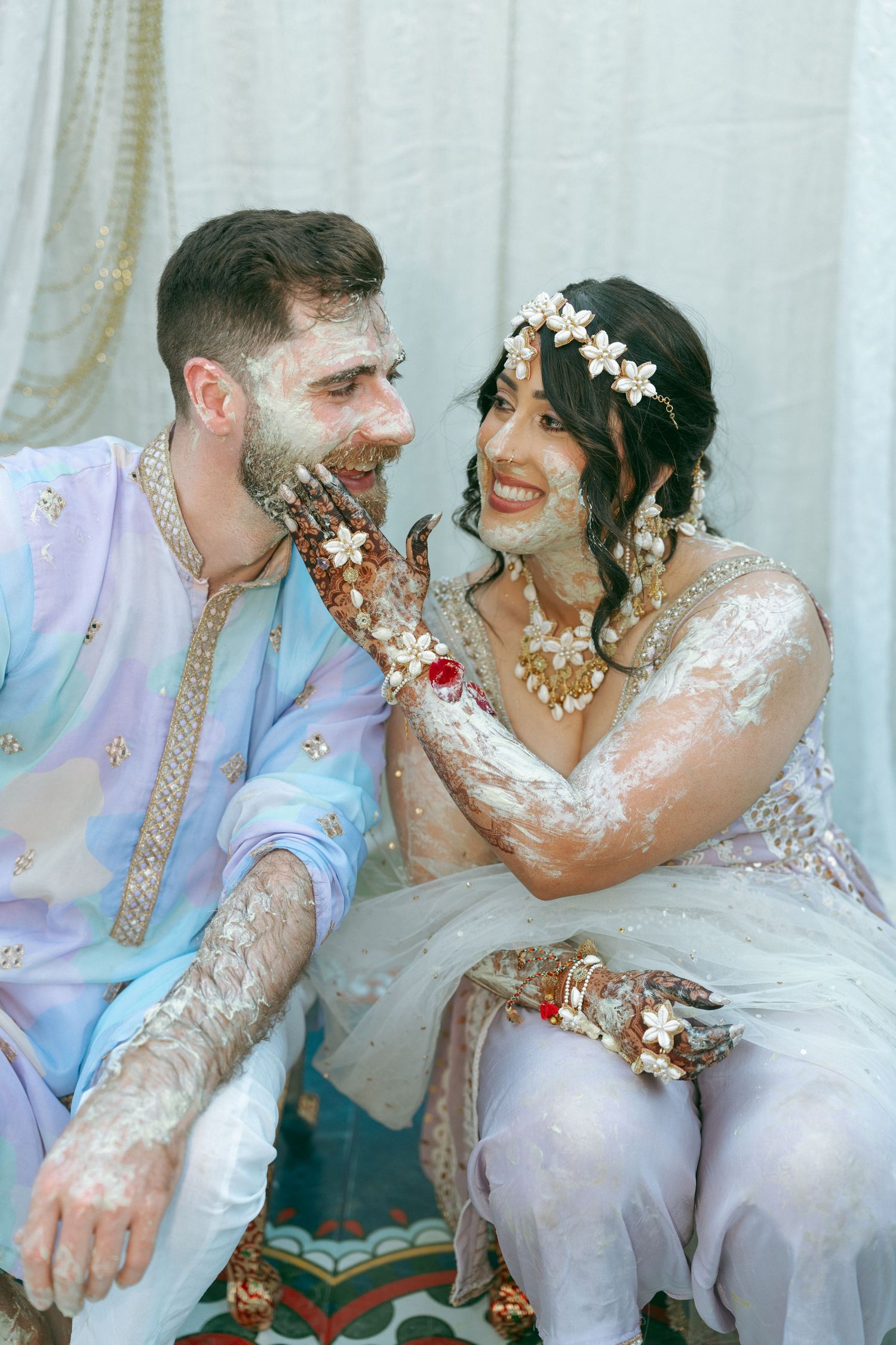 Couple in traditional Indian attire laughing during Haldi ceremony, covered in white paste, wearing floral accessories.