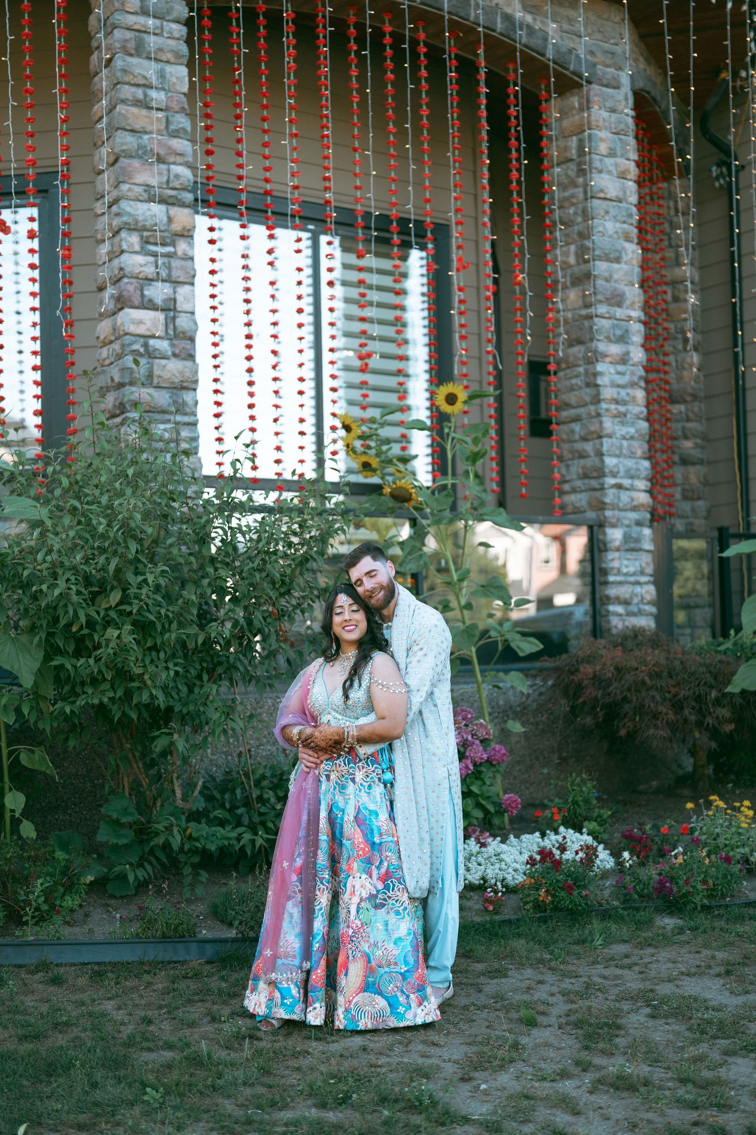 Couple in traditional Indian attire posing in a garden with decorative floral hangings and stone building backdrop.