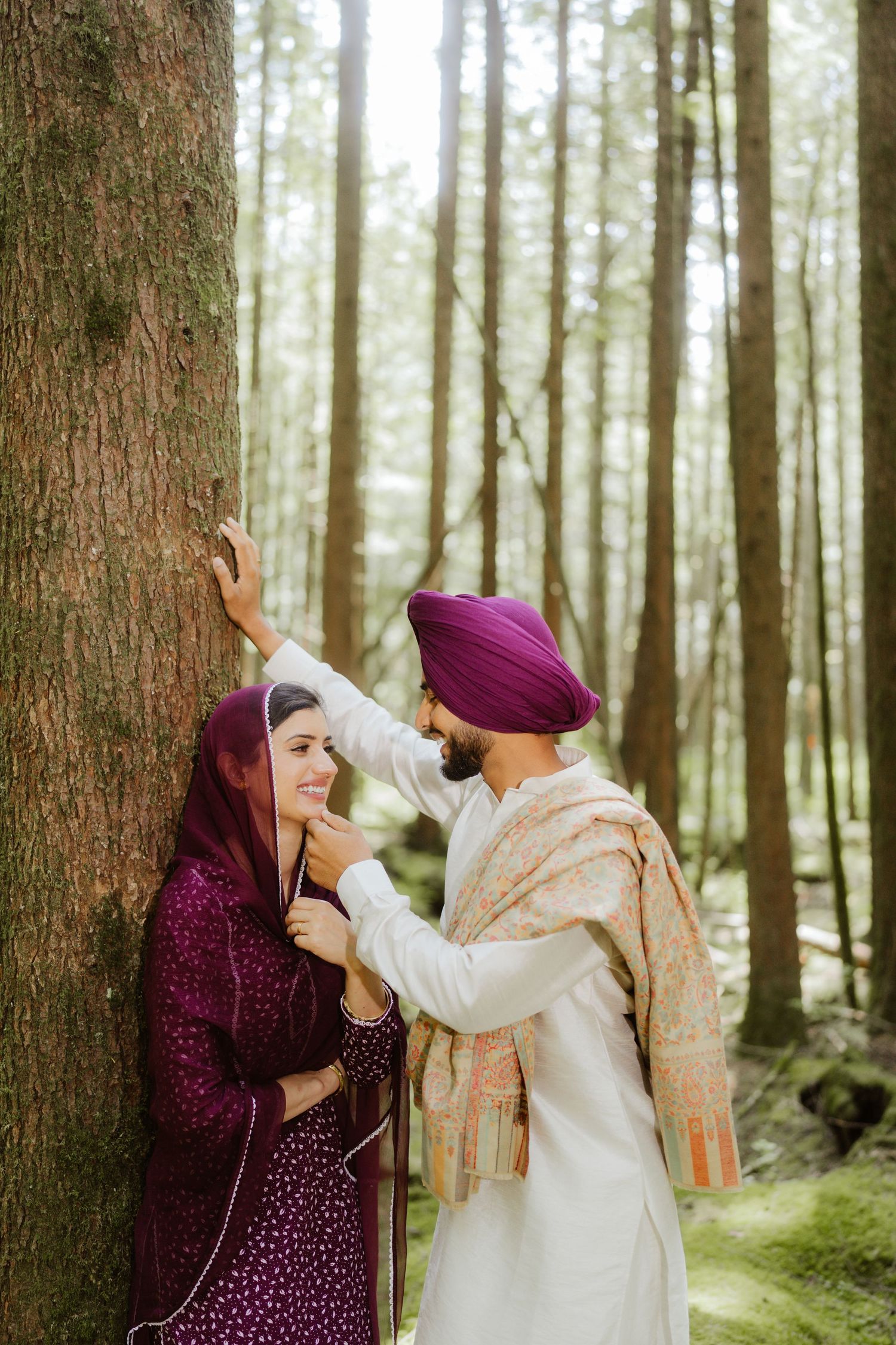 Sikh couple sharing a romantic moment by a tree in a forest, man in purple turban adjusting womans dupatta.