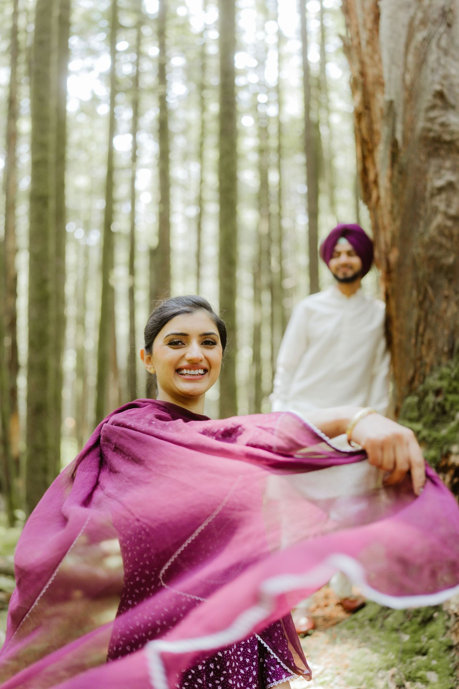 Smiling South Asian woman in burgundy salwar suit twirling dupatta with man in white kurta in forest.