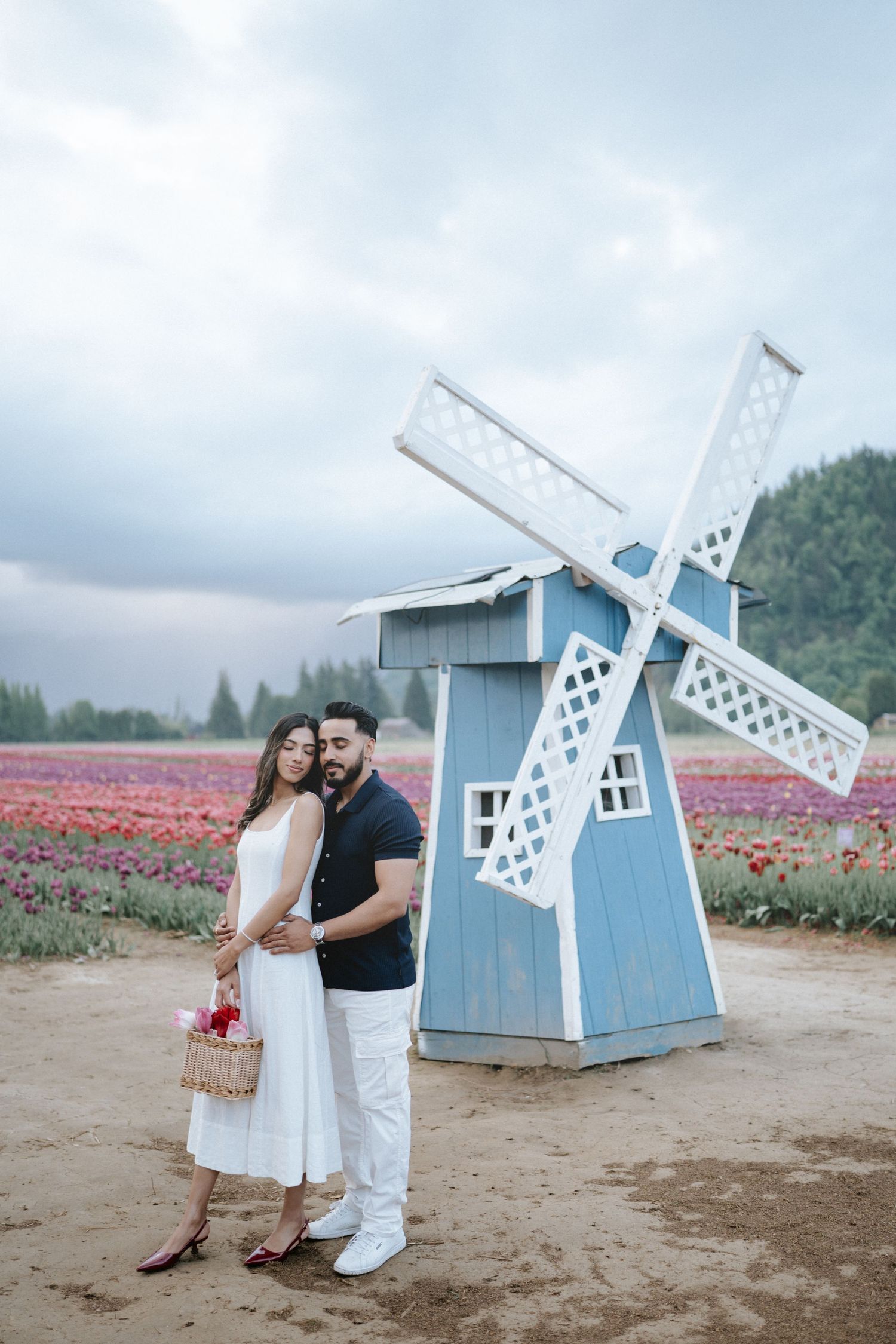 A couple poses near a decorative blue windmill structure in a vibrant tulip field on an overcast day.