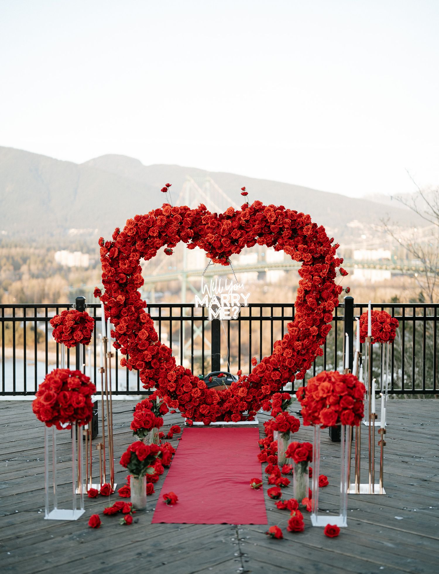 A romantic outdoor wedding ceremony setup with red rose arches and a pink carpet overlooking a mountain landscape.