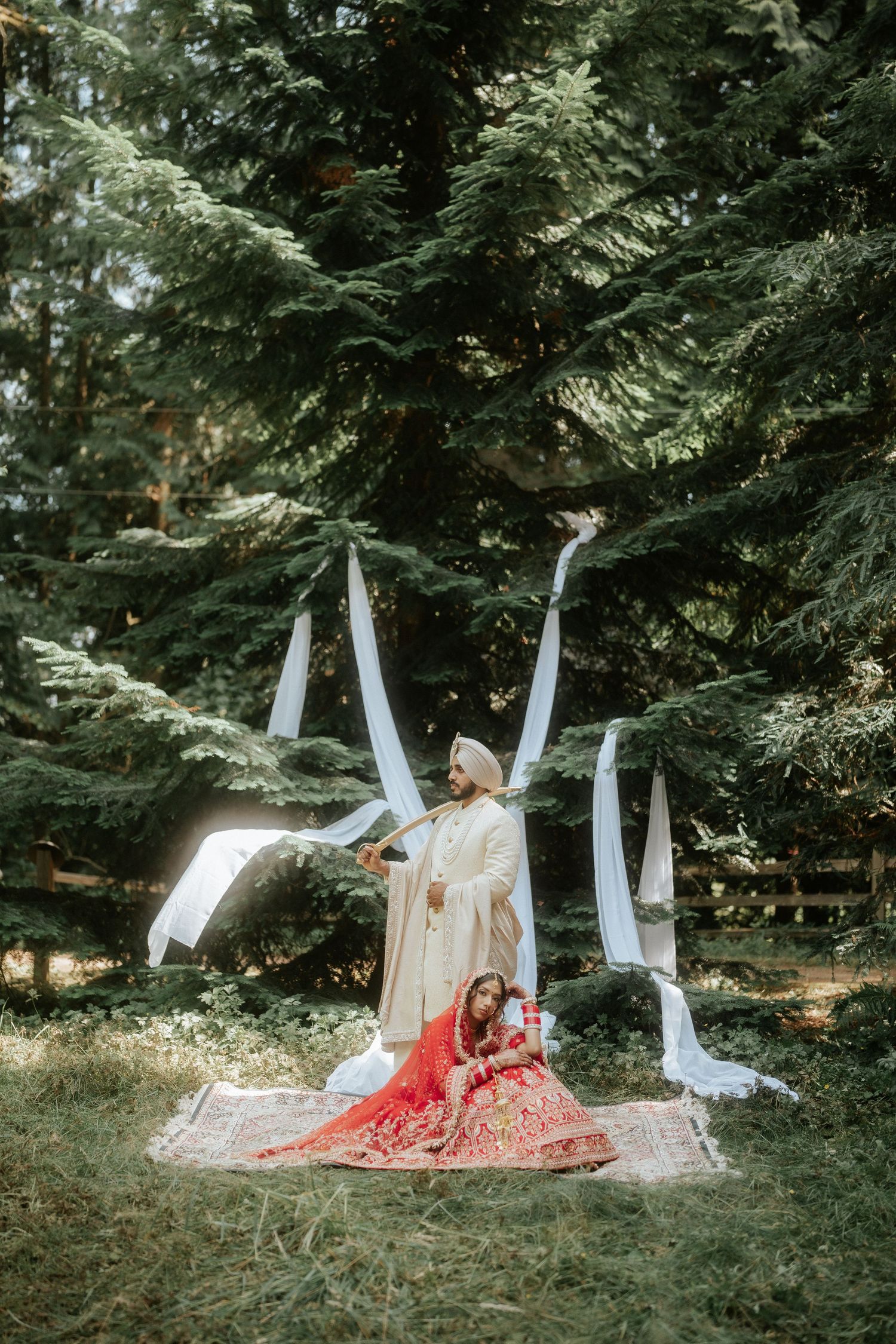 A wedding couple poses on the ground with white fabric draping in a wooded outdoor setting.