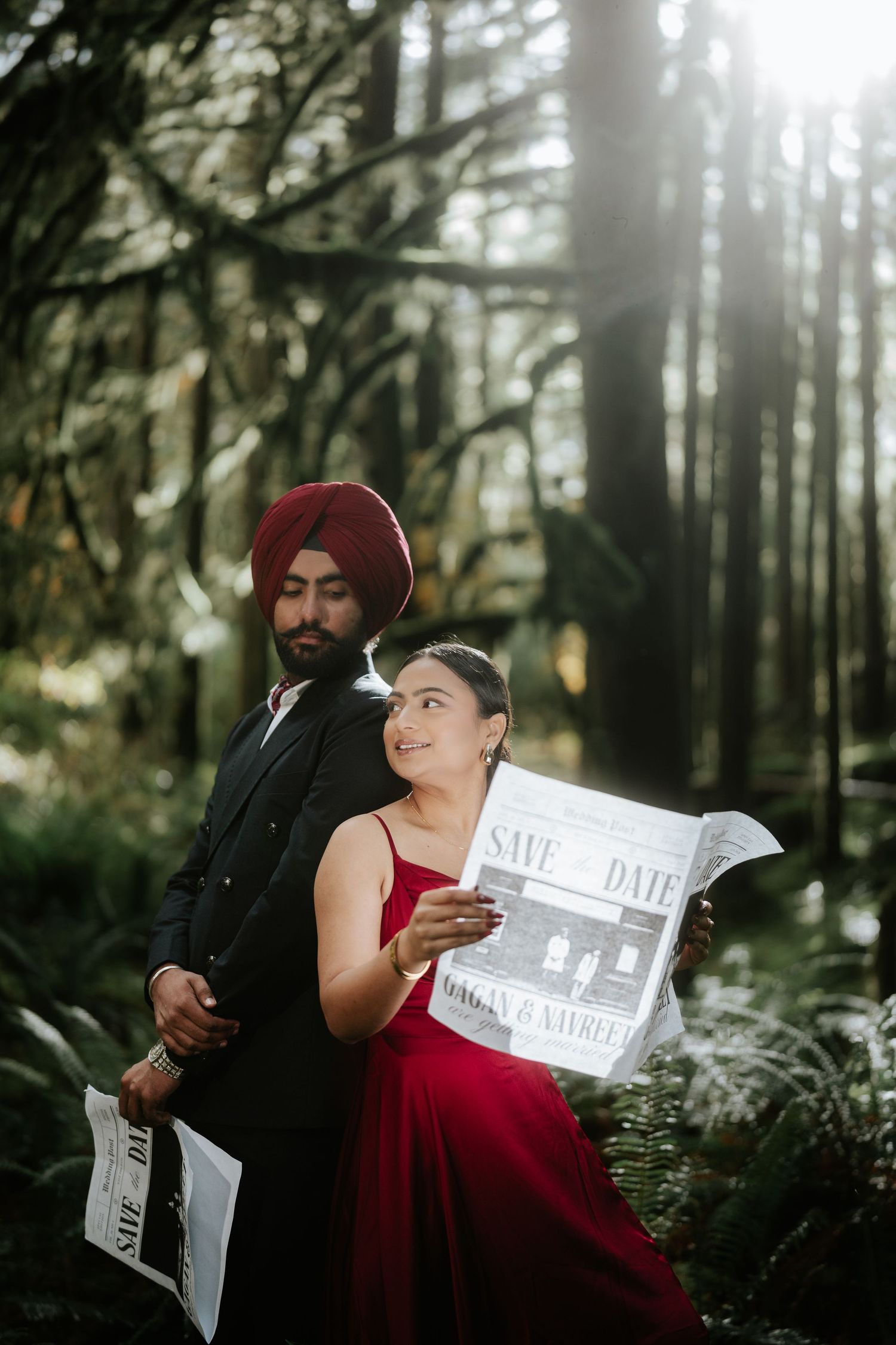 A stylish couple poses with save-the-date newspapers in a moody forest setting.