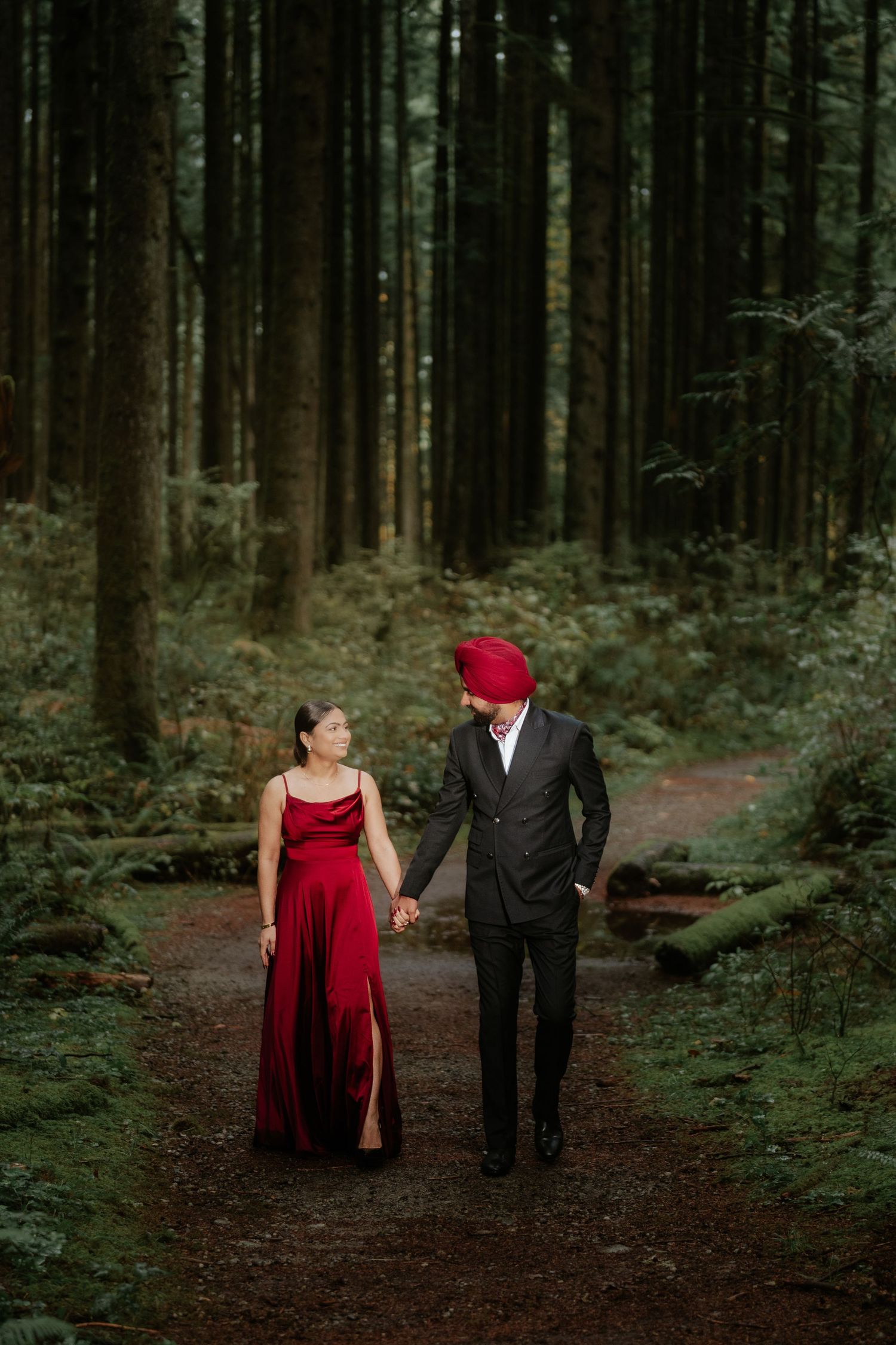 Couple in red dress and black suit walks through a moody forest setting.