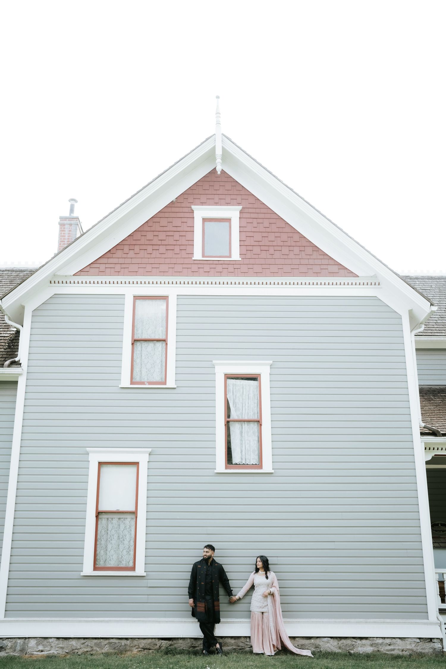 A couple poses against pale blue historic houses with white trim.