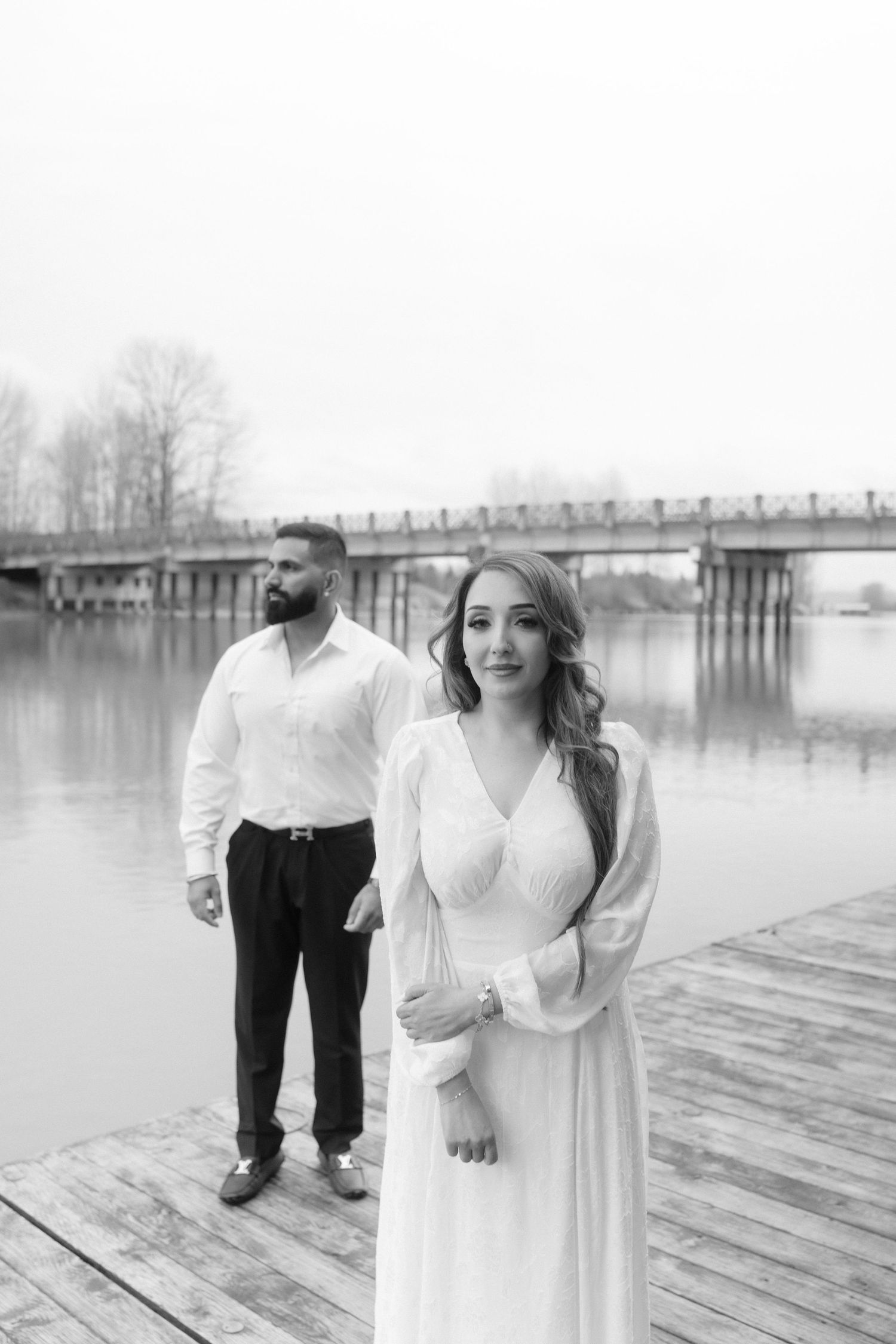 A black and white portrait of a couple on a wooden dock with a bridge in the background.