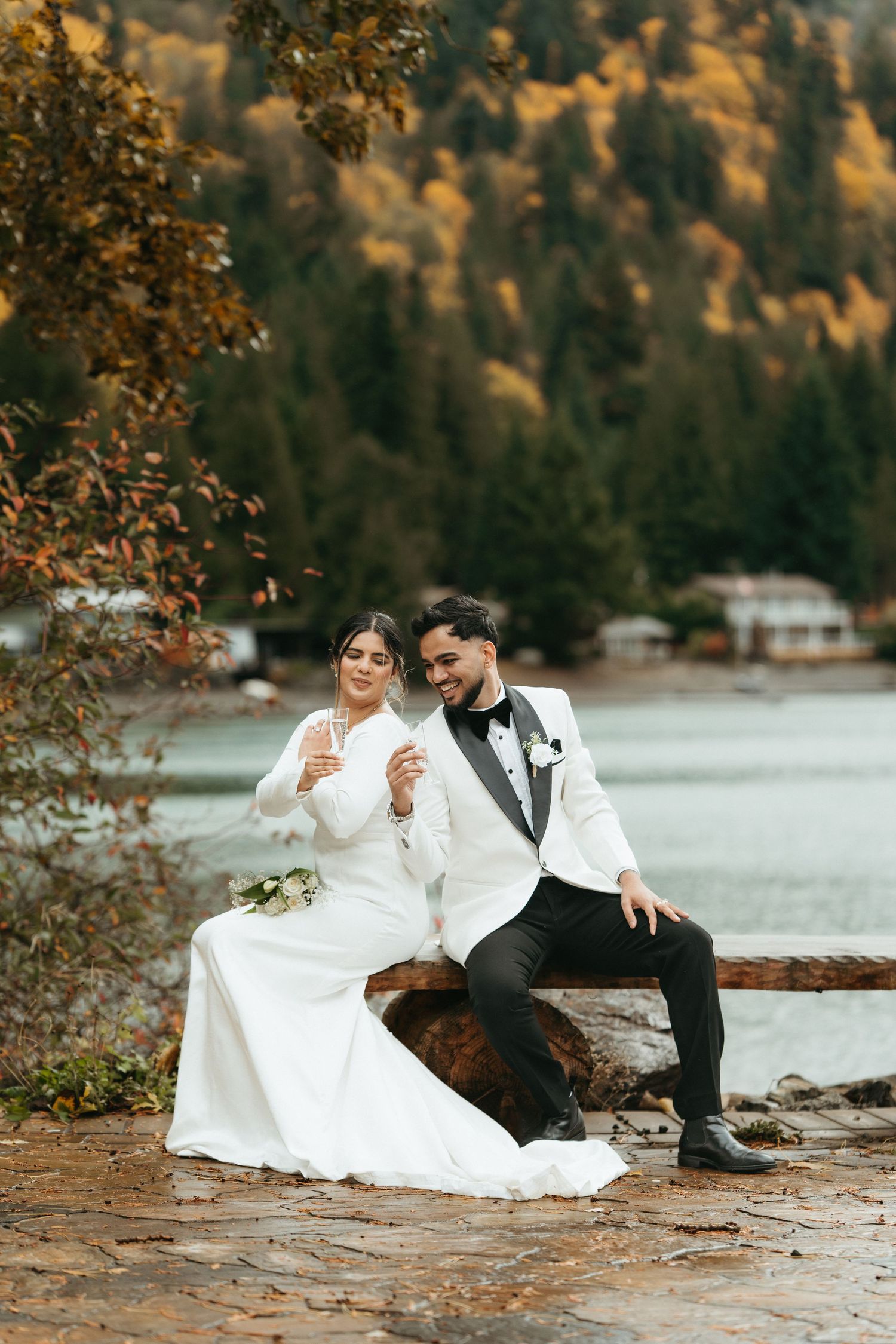 A couple in wedding attire sits by a lakeside during autumn with mountains and fall foliage in the background.