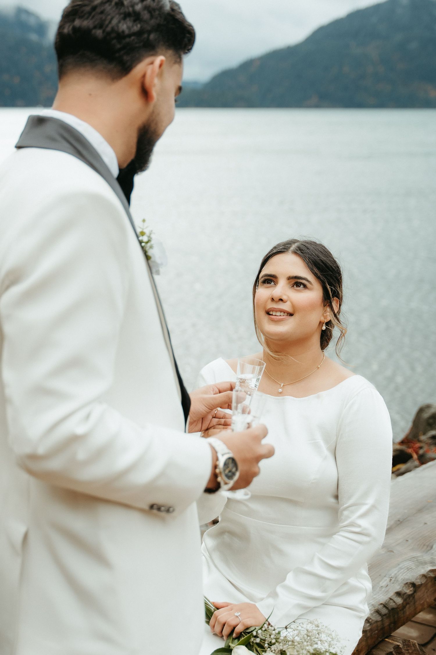 Couple in white wedding attire sharing intimate moment by misty mountain lake during outdoor ceremony.