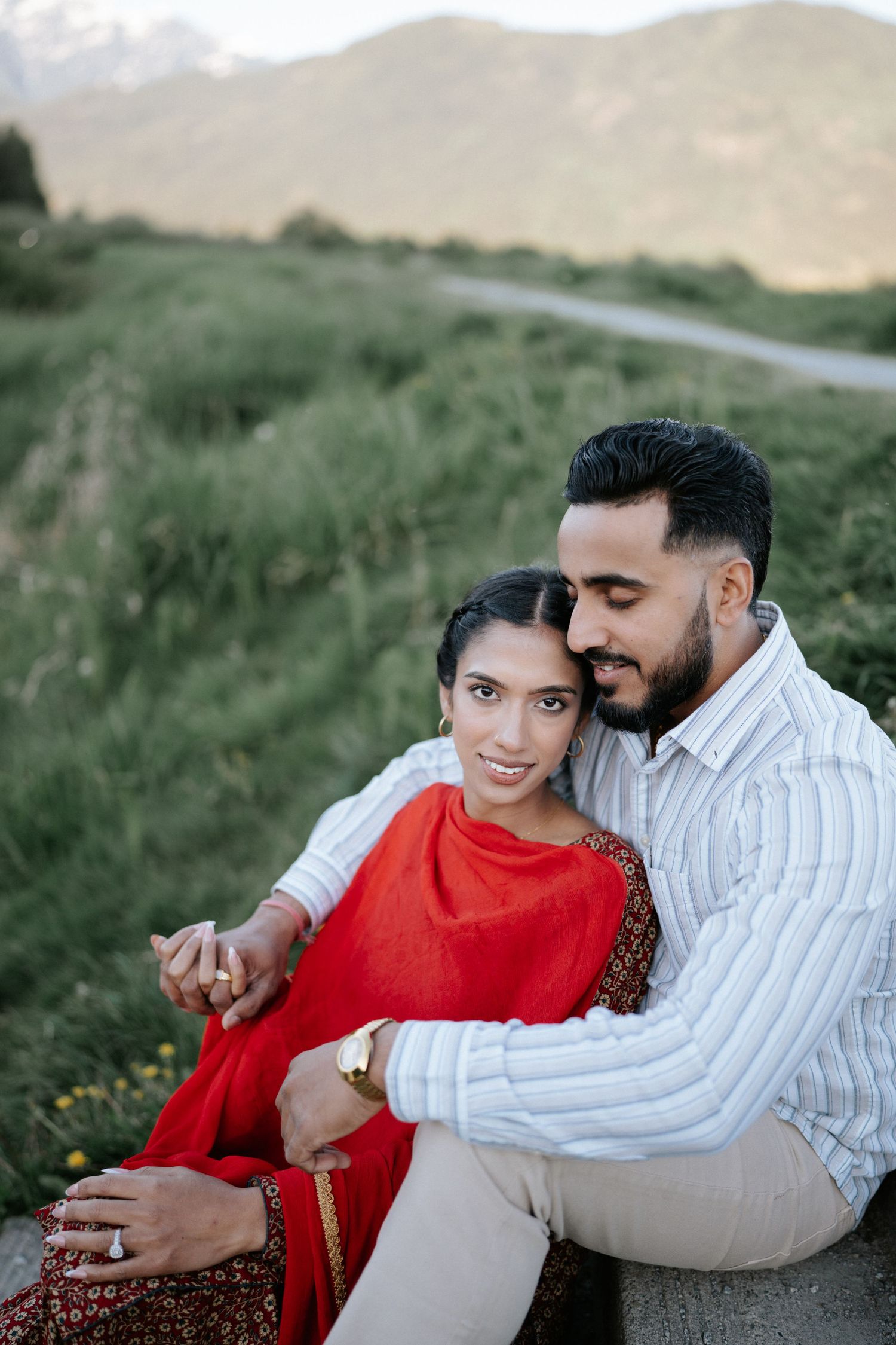 A couple in red and white outfits share intimate moments while posing together in a grassy outdoor setting at sunset.