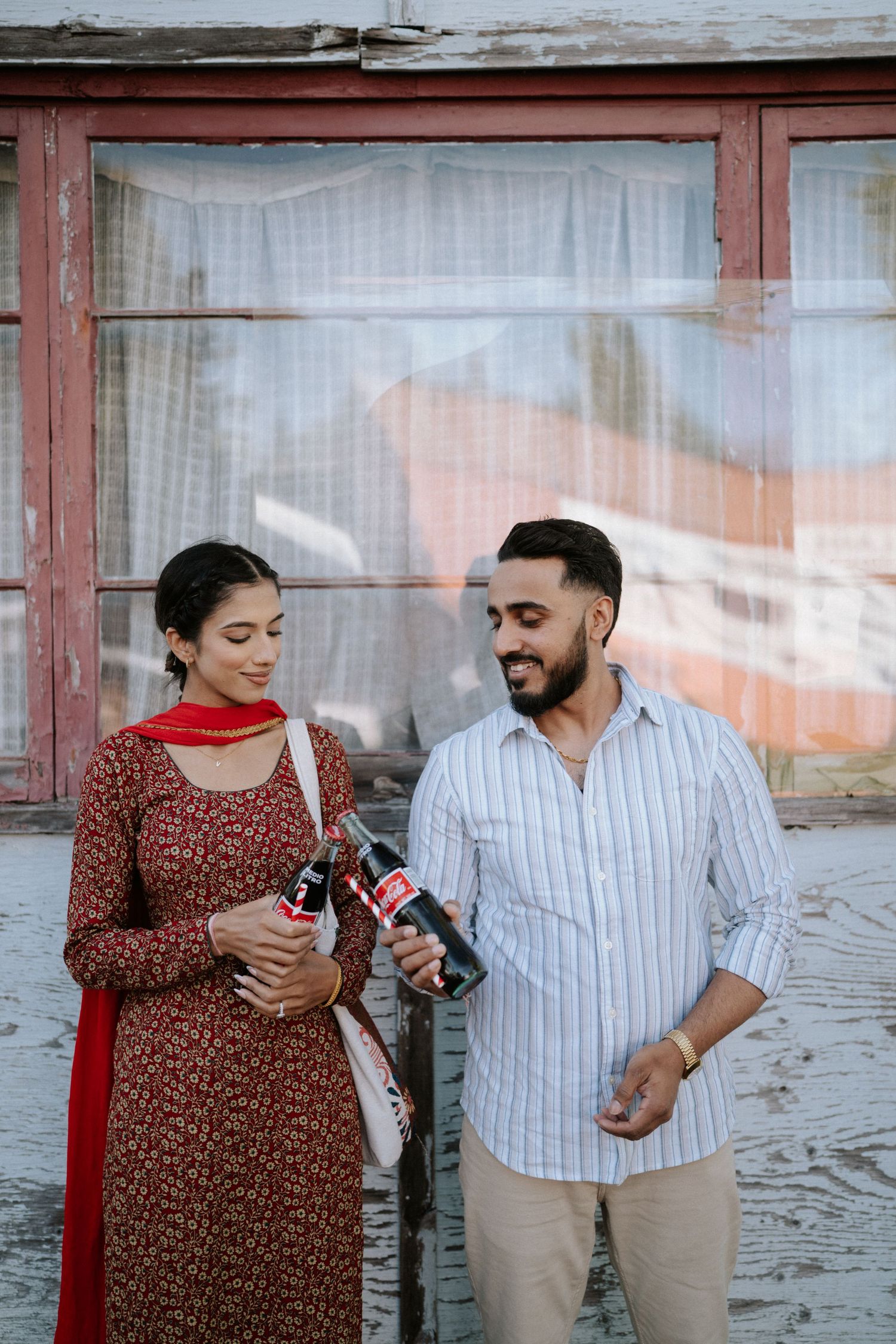 A couple sharing a happy moment while holding Coca-Cola bottles against a rustic wall with window backdrop.