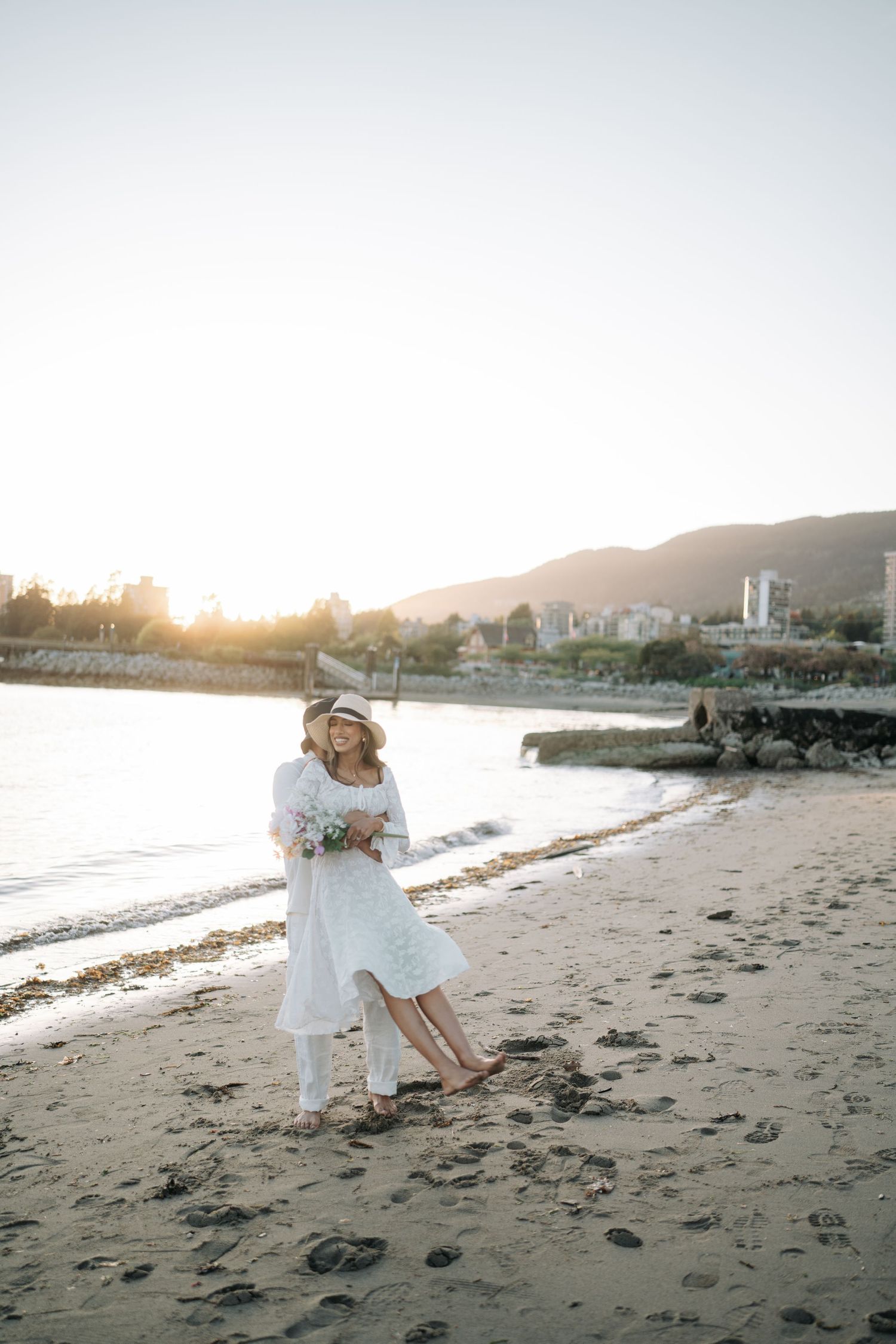 Couple embraces on a sandy beach at sunset with coastal buildings in the background.