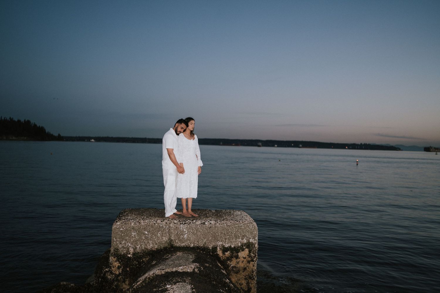 A couple stands together on a rocky ocean overlook during sunset while holding a white fabric in the breeze.
