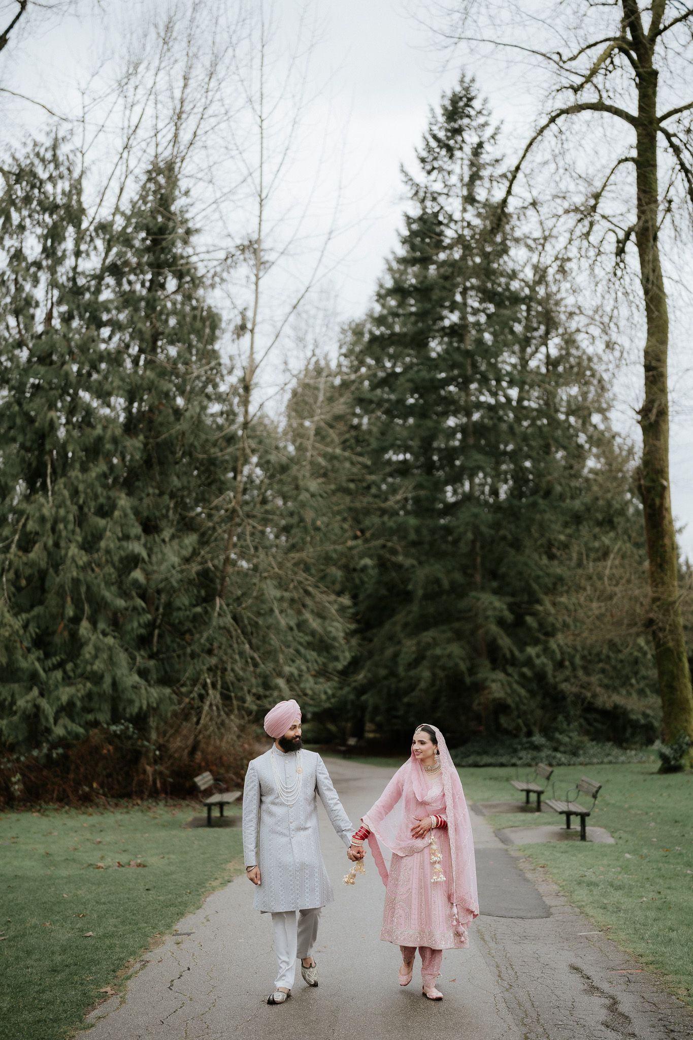 A couple walks hand in hand along a tree-lined path in a park wearing traditional South Asian wedding attire.