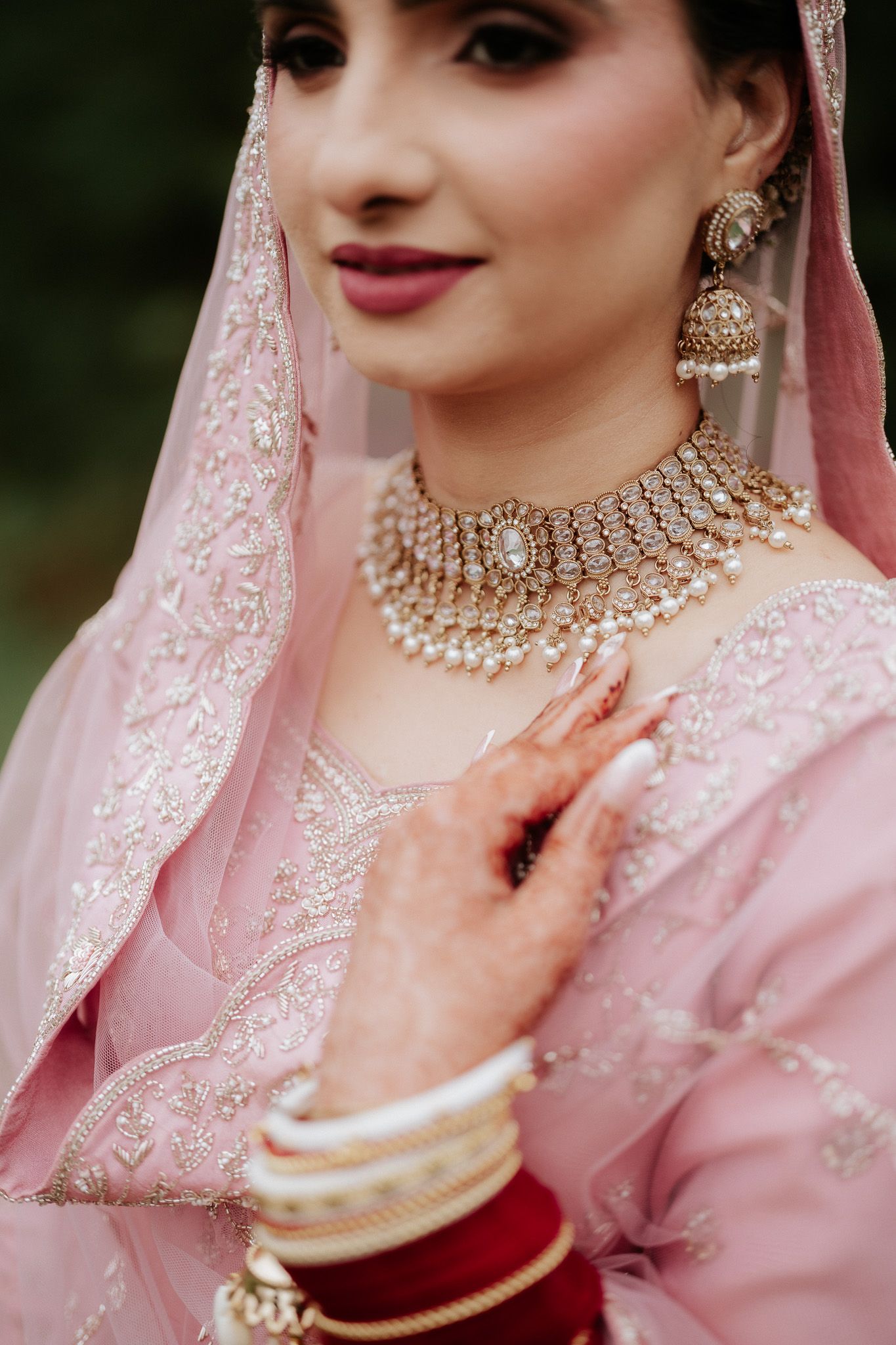 Traditional South Asian bride wearing a pink embroidered outfit with pearl jewelry and intricate neckpiece details.
