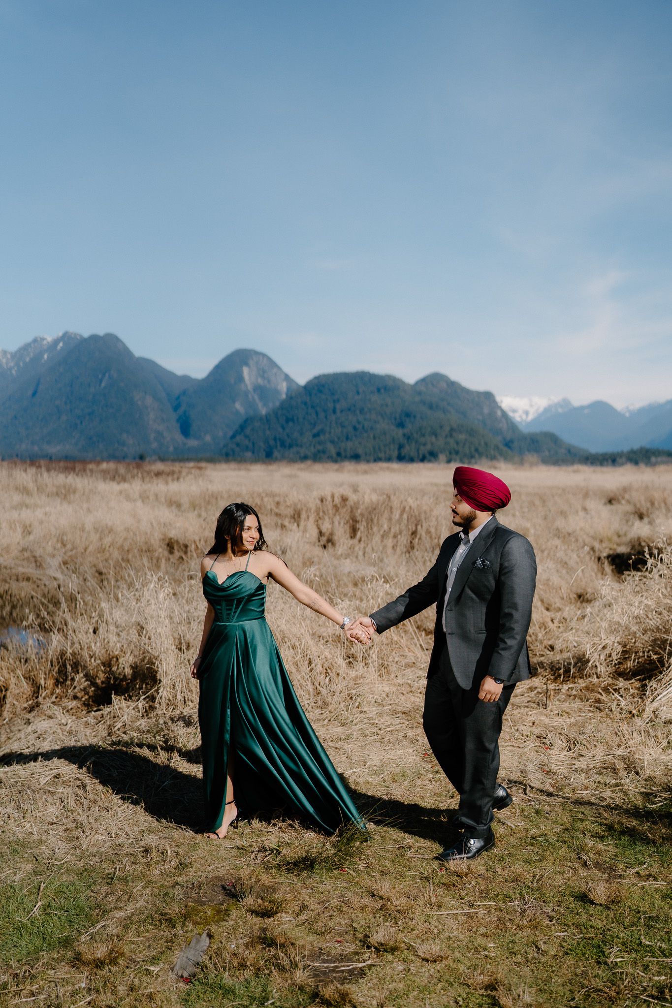 Couple in formal attire dance and pose together in a scenic mountain meadow with golden grass and mountain peaks behind them.
