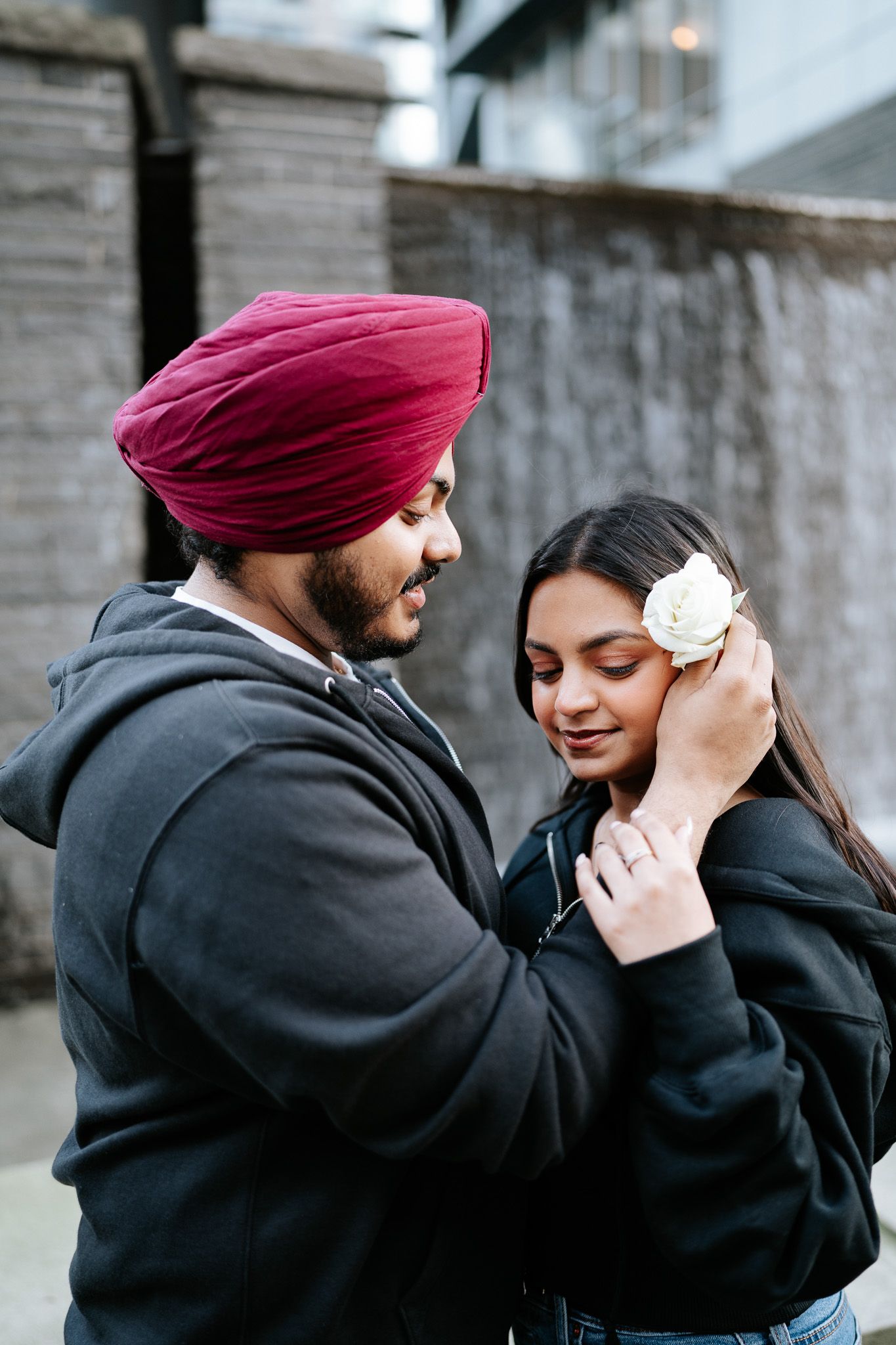 A couple in traditional Sikh attire share an intimate moment against a stone wall background.