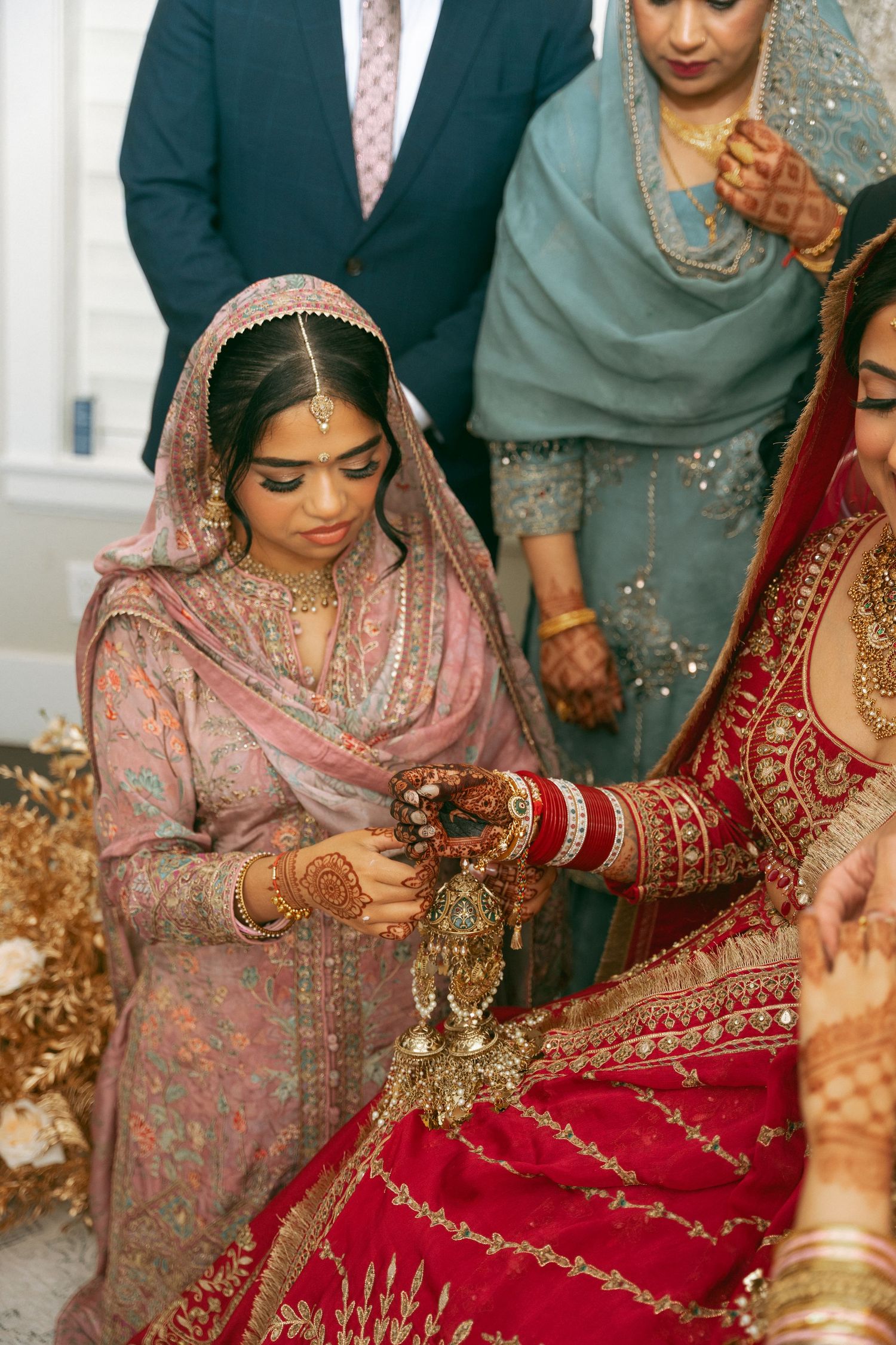 South Asian bride in red bridal attire with gold jewelry during traditional wedding ceremony with women in elegant dress.