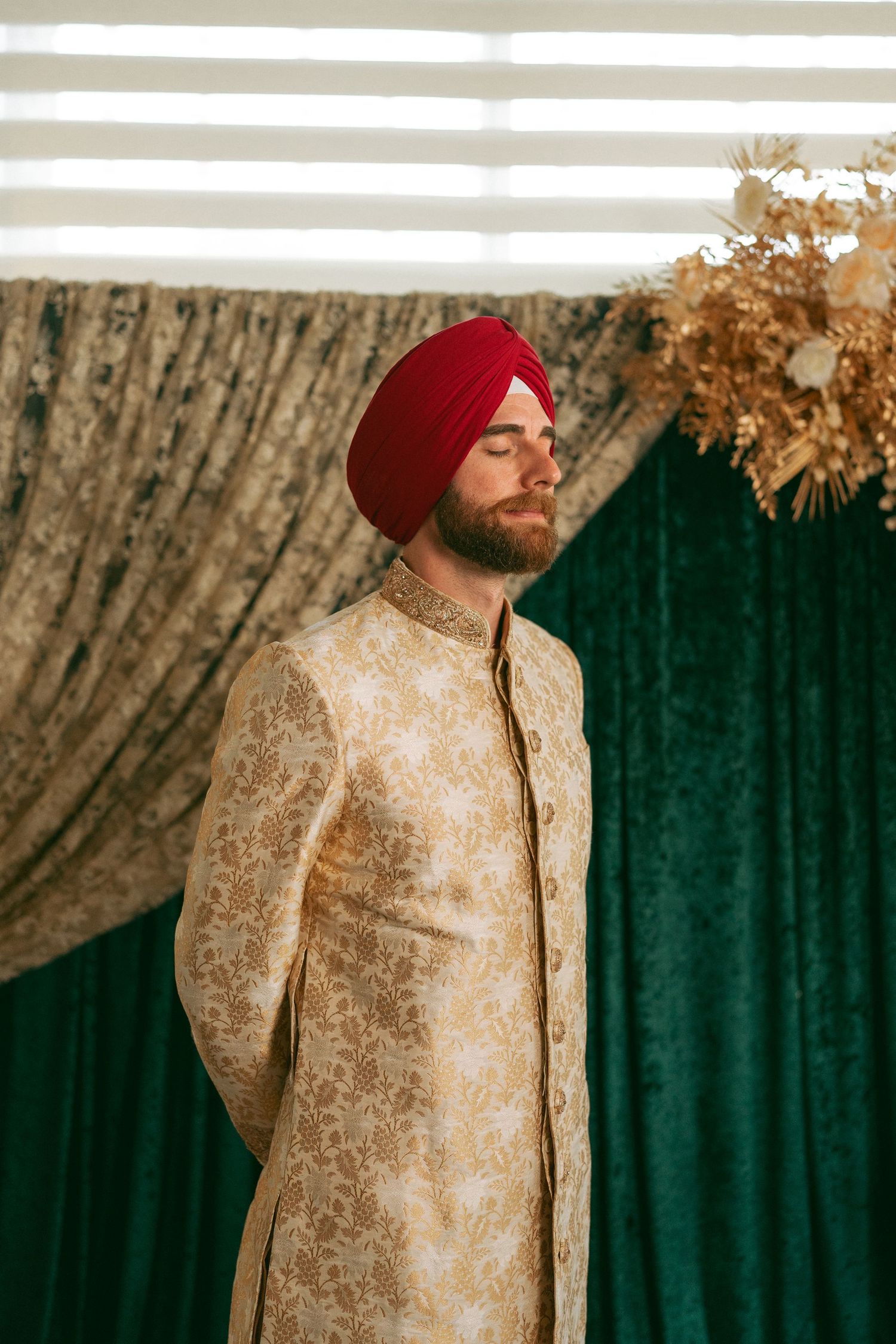 Man in gold sherwani and red turban stands with eyes closed before green velvet curtain backdrop.