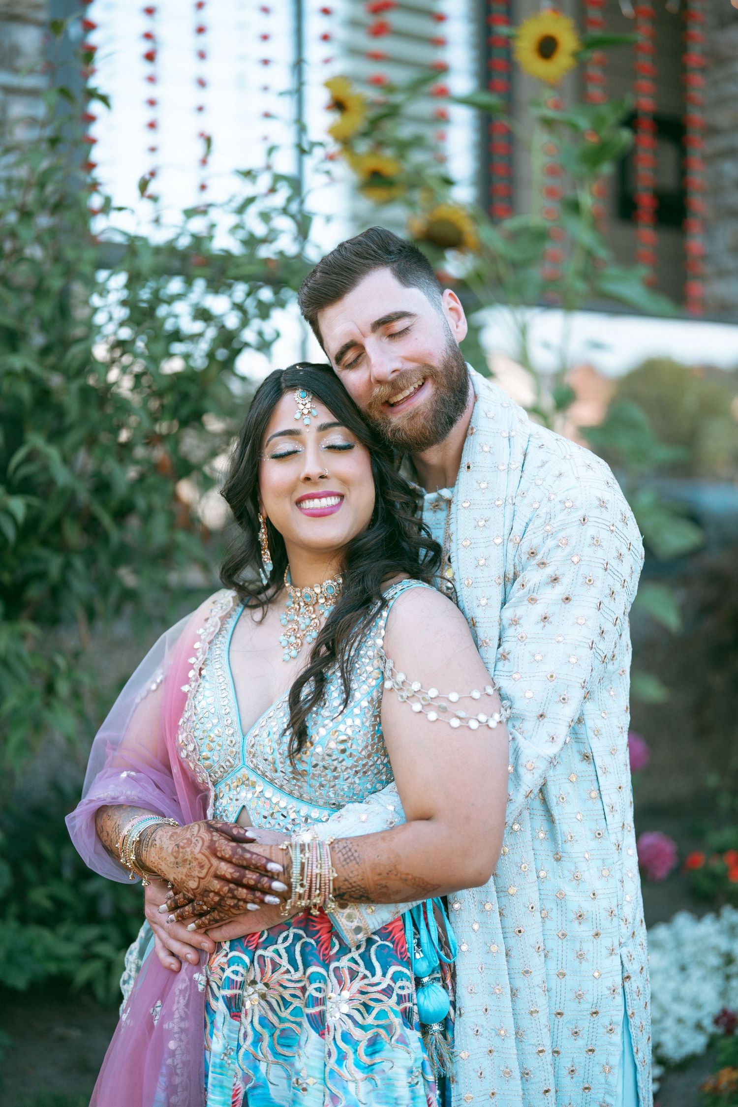 Couple in traditional South Asian wedding attire embrace outdoors, bride in blue lehenga with mehndi, groom in sherwani.