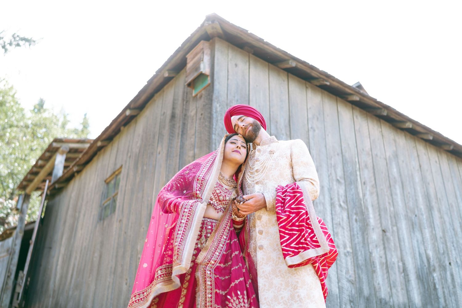 South Asian couple in traditional pink and gold wedding attire embracing outdoors near rustic wooden barn.