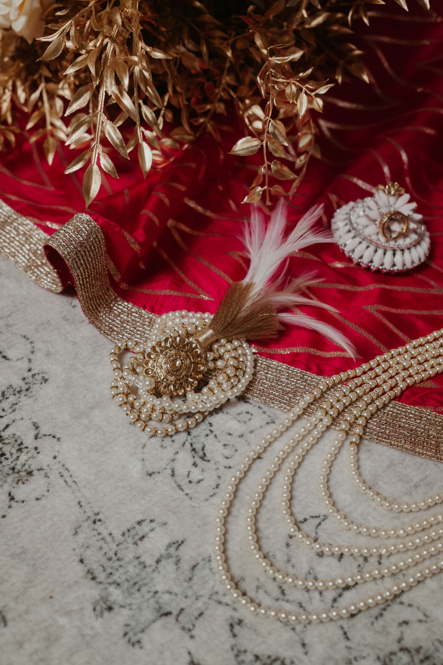 Elegant bridal accessories with pearl necklace, gold belt, feathers, and rings on red fabric backdrop.