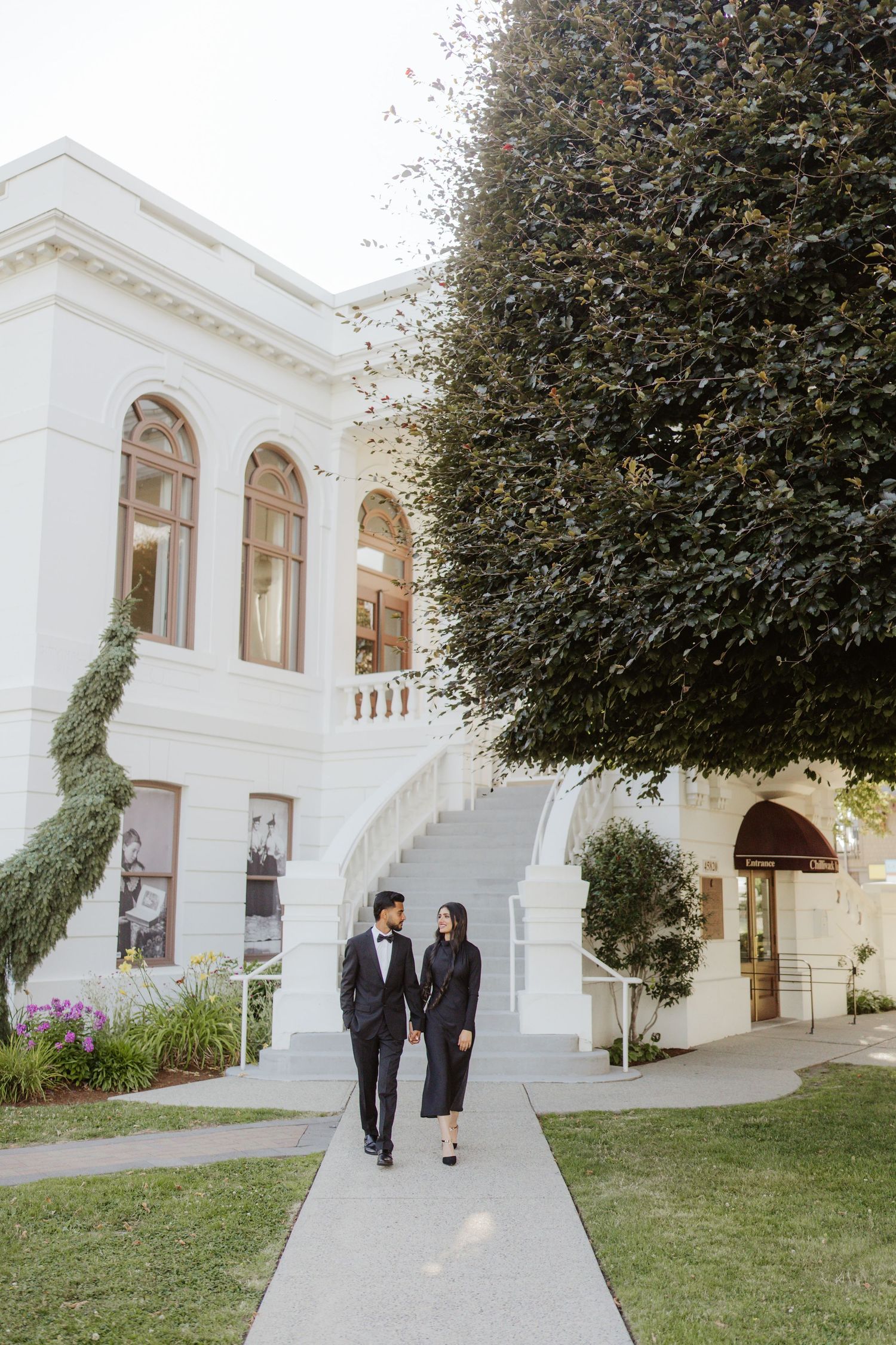 Elegant couple in matching black outfits walking along pathway of white colonial building with large tree.
