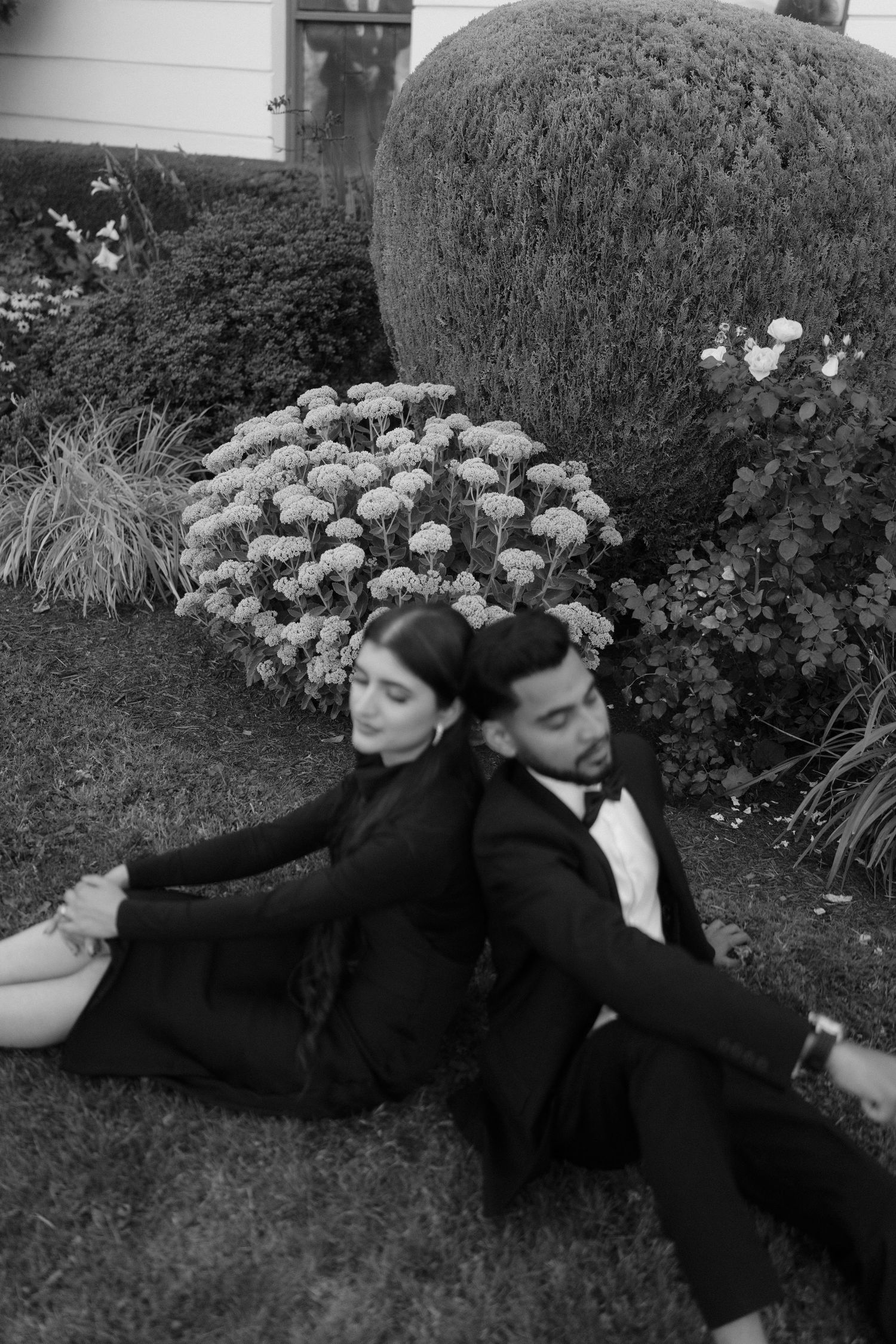 Black and white photo of a couple in formal attire sitting back-to-back on grass in a garden.